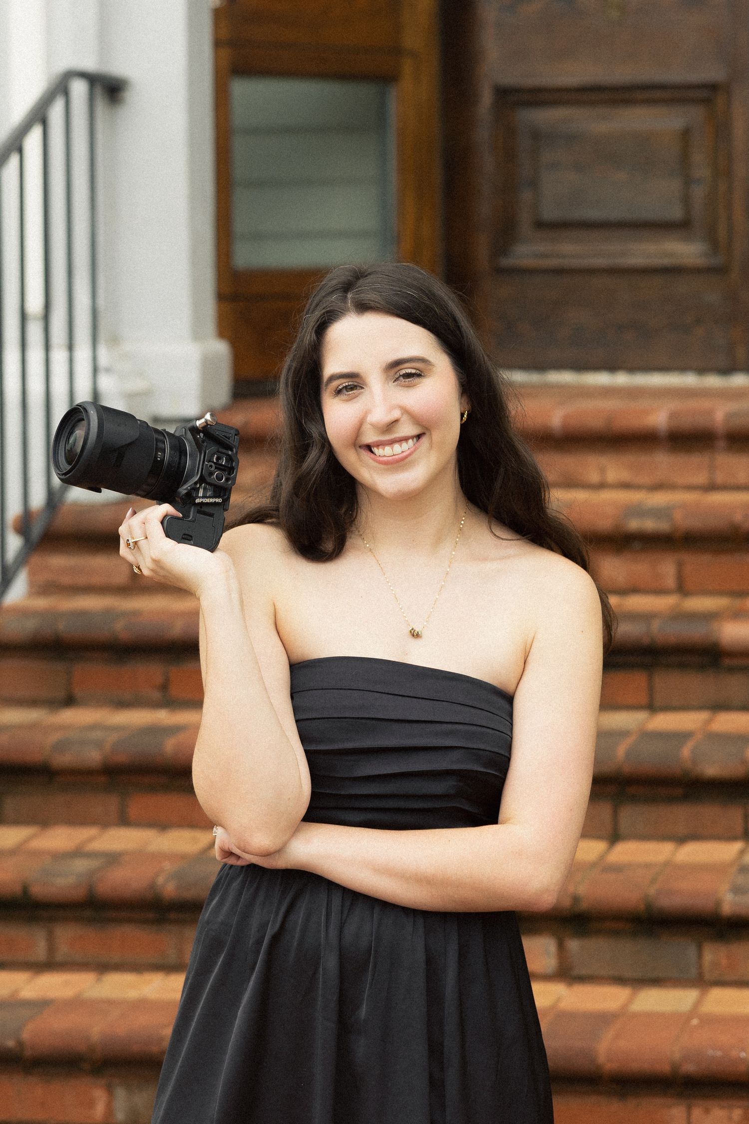 Smiling woman in black strapless dress holding a DSLR camera on brick steps.