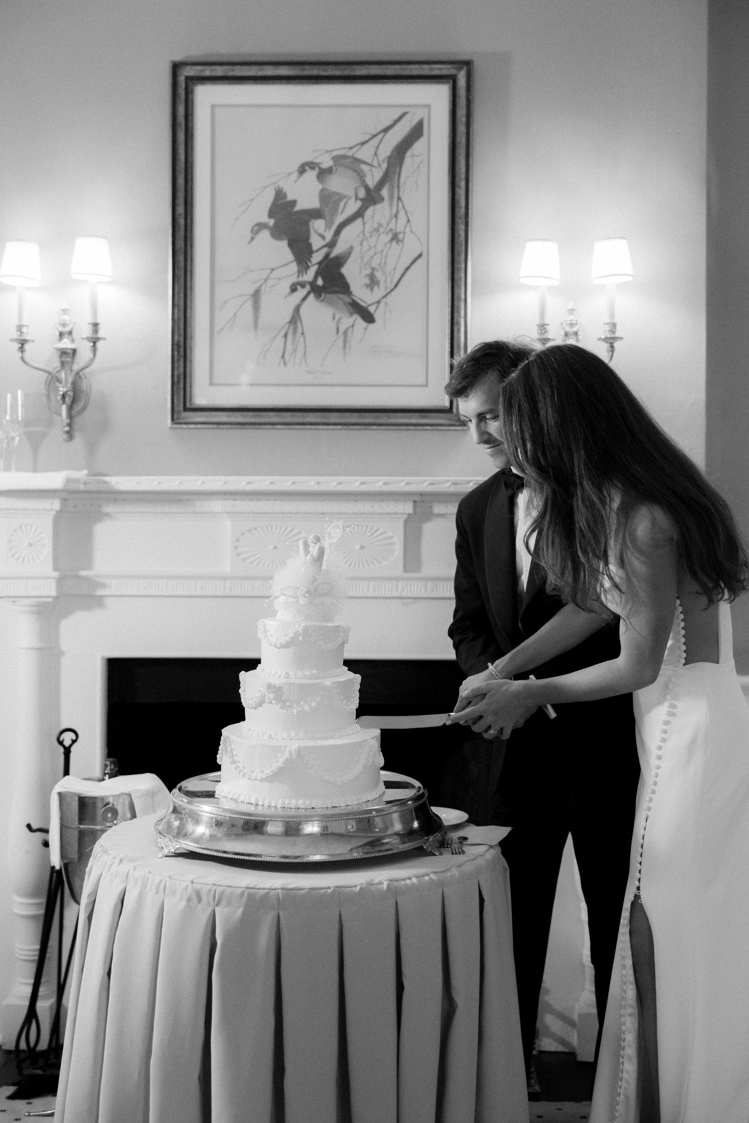 Black and white photo of bride and groom cutting their elegant four-tier wedding cake near a fireplace.