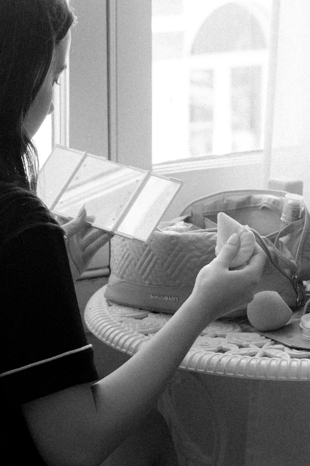 Black and white photo of a woman sitting by a window, examining items on a wicker table with a designer bag.