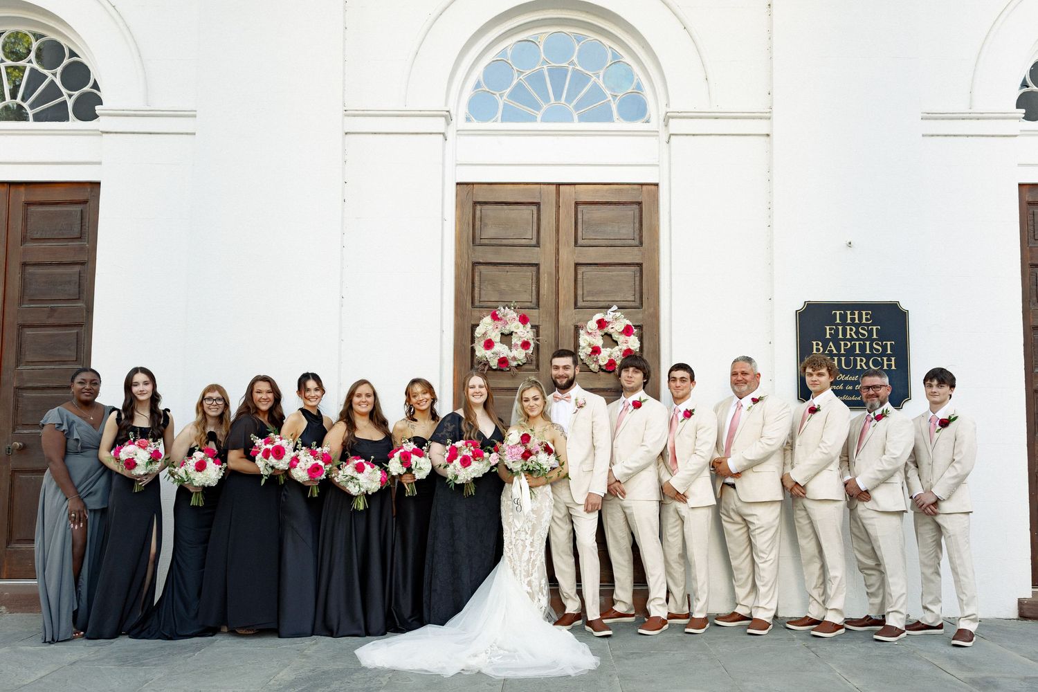 Wedding party poses outside First Baptist Church, bridesmaids in black gowns and groomsmen in tan suits holding pink floral bouquets.
