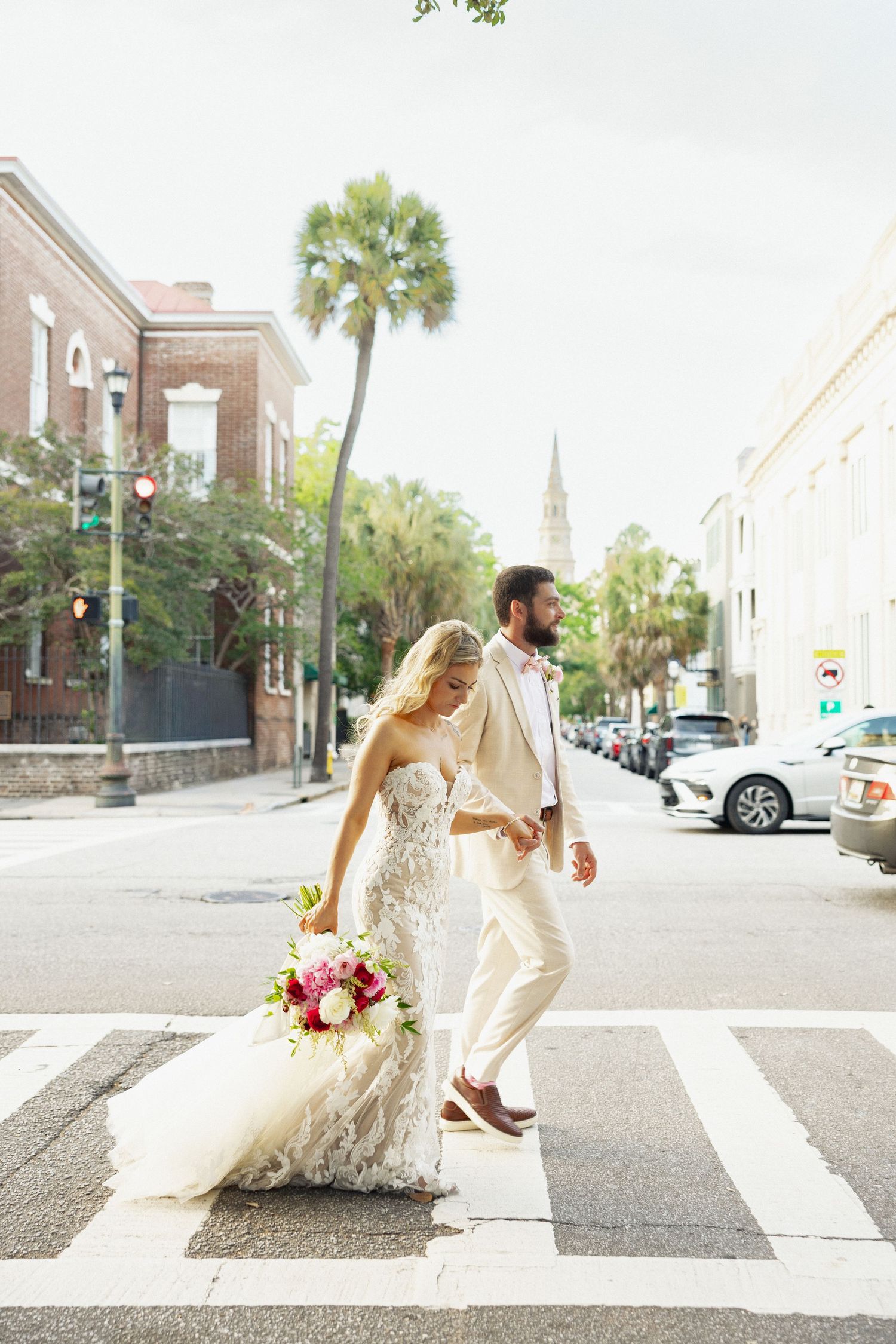 Bride in lace gown with vibrant bouquet and groom in cream suit cross sunny Charleston street with palm trees.