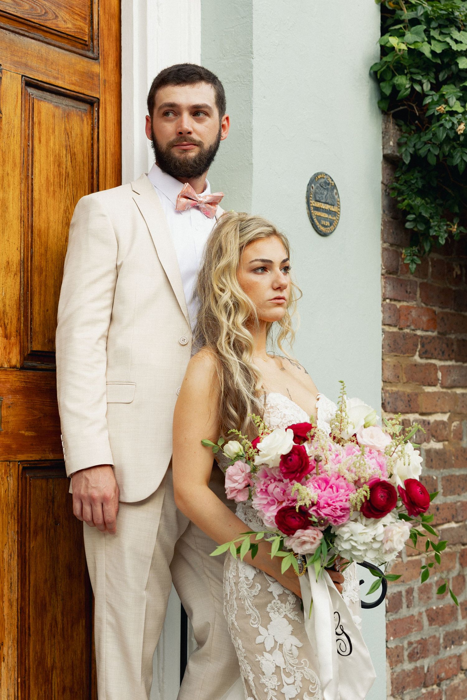 Bride and groom posing by historic Charleston doorway, lace wedding gown, tan suit, pink bow tie, vibrant floral bouquet.