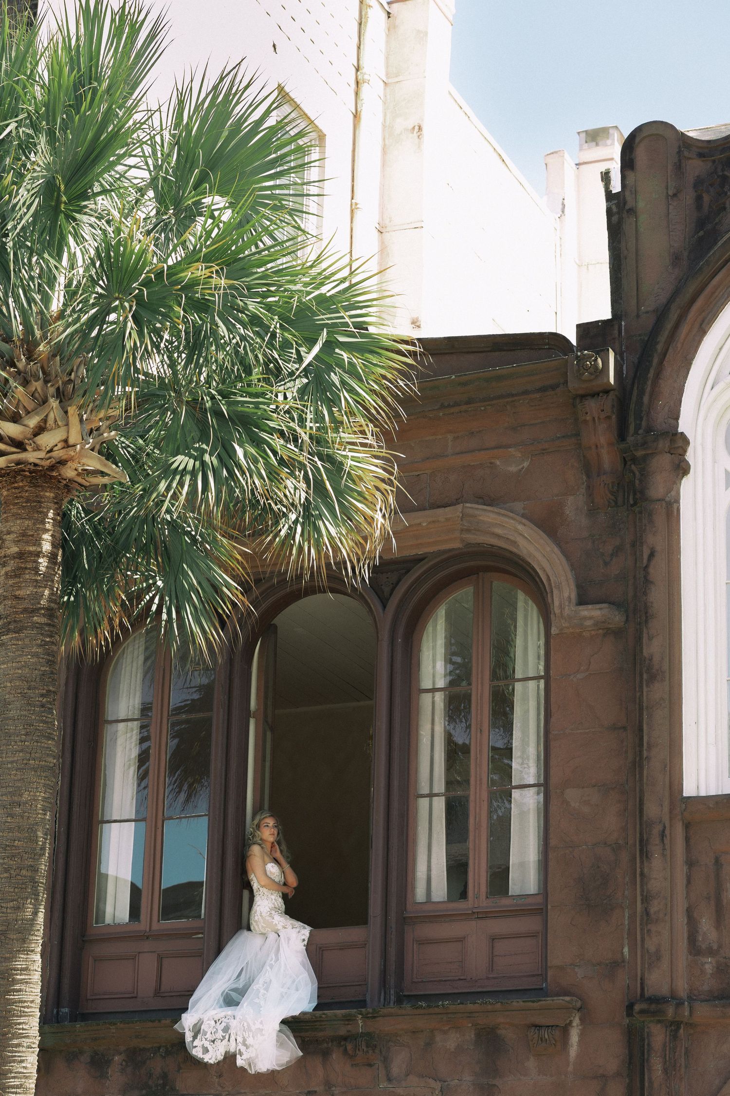 Bride in white wedding gown standing on ledge of ornate brick building with arched windows and palm tree.