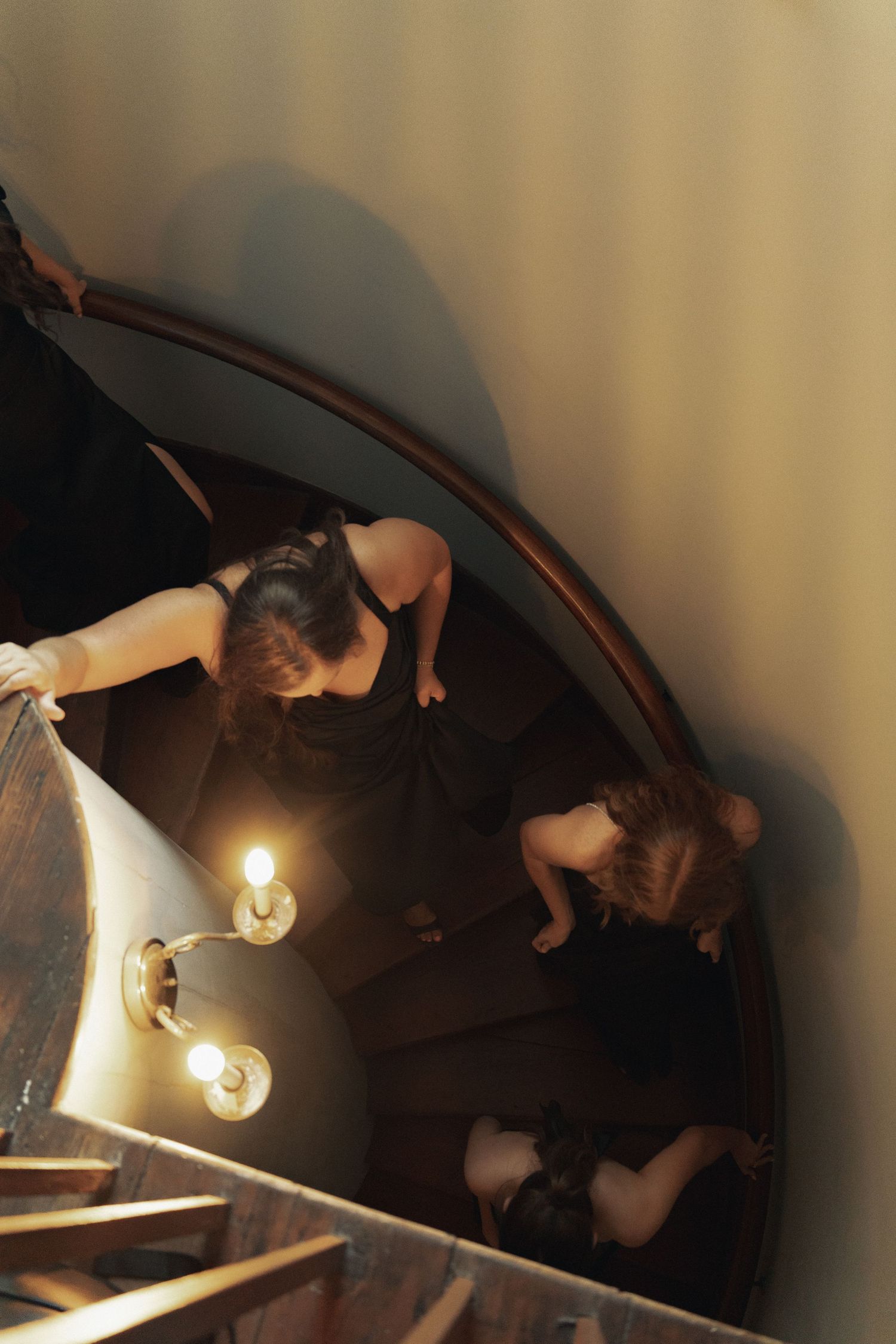 Aerial view of two women in black dresses descending an elegant spiral staircase with brass wall lights.