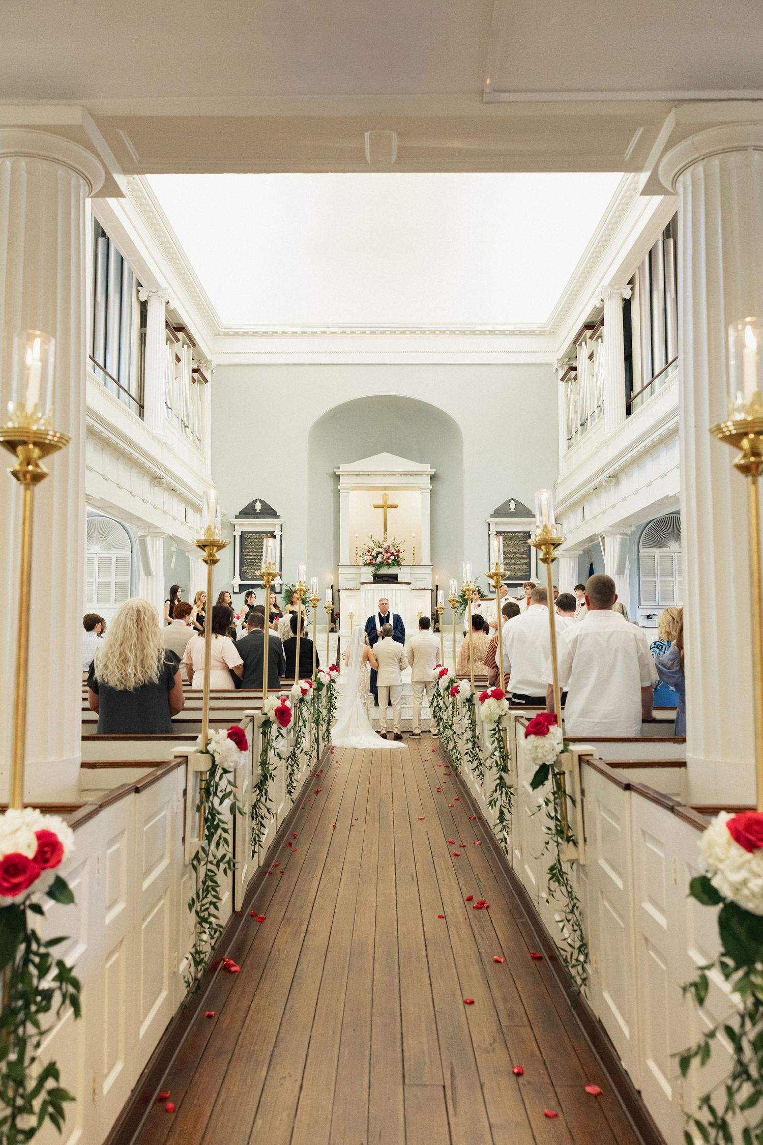 Wide view of elegant church wedding ceremony with rose-adorned pews and guests seated along wooden aisle.