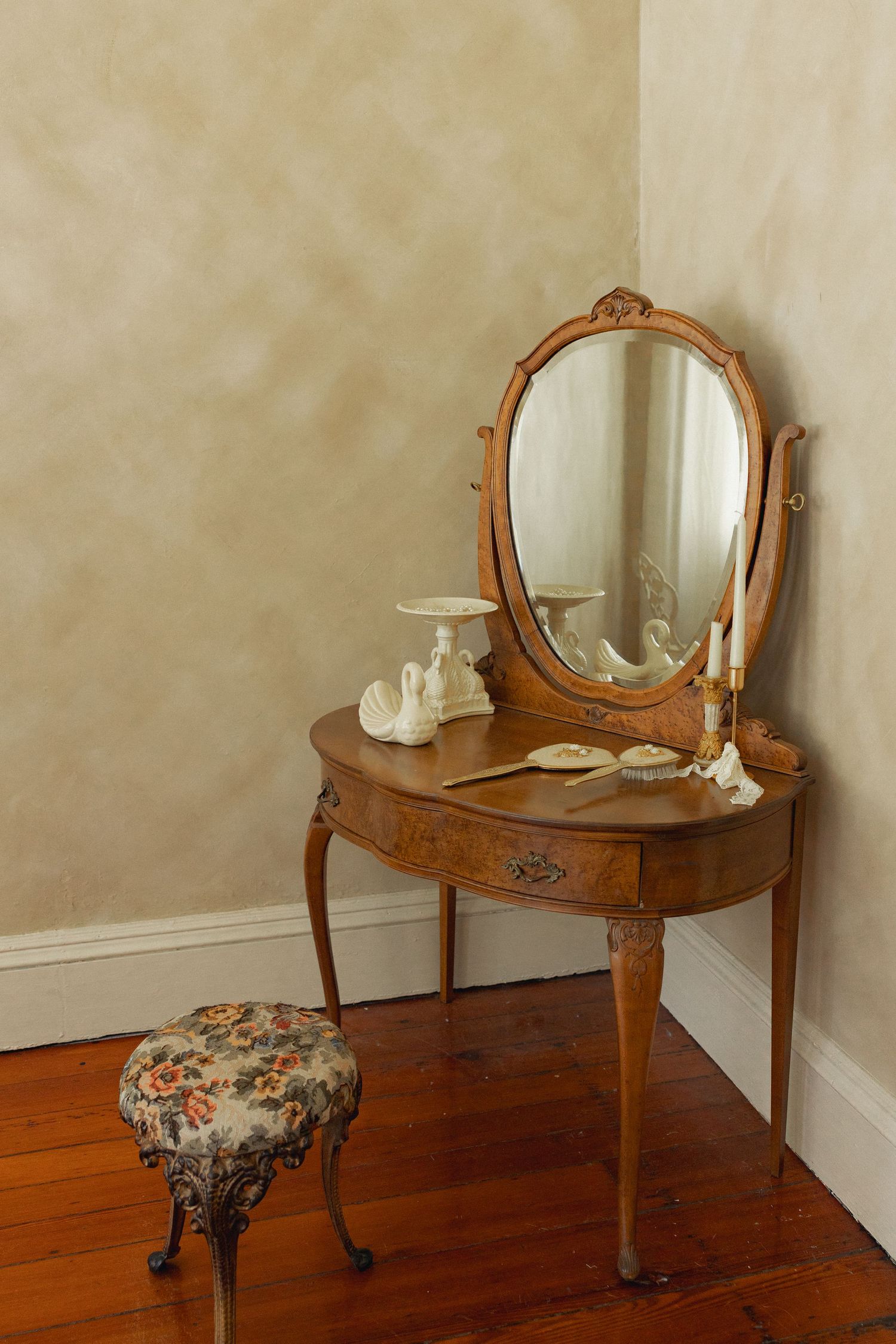 Antique wooden vanity with oval mirror, floral stool, and decorative items on hardwood floor in corner.