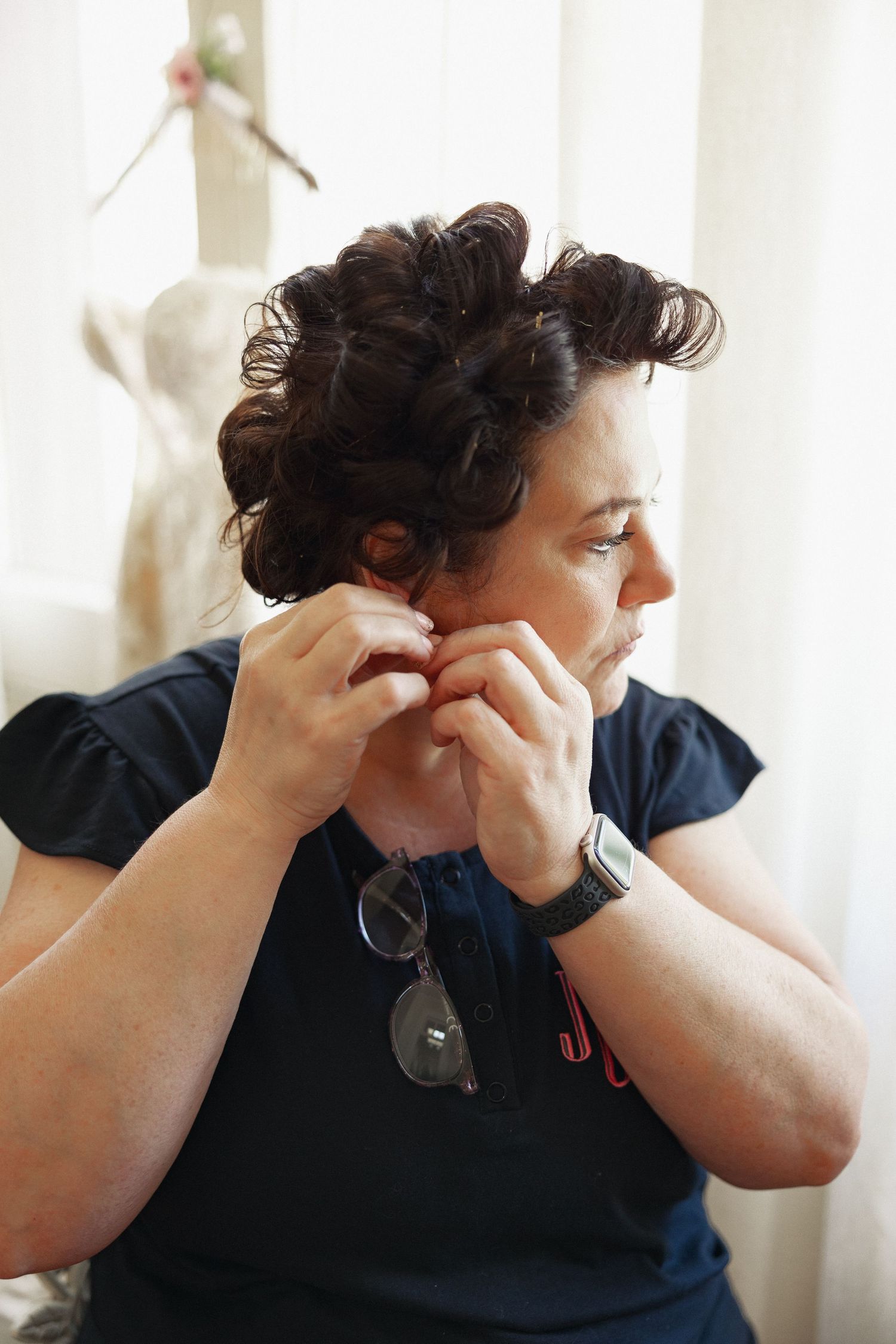 Woman with curly dark hair putting on earrings wearing navy blue t-shirt with sunglasses and silver watch.