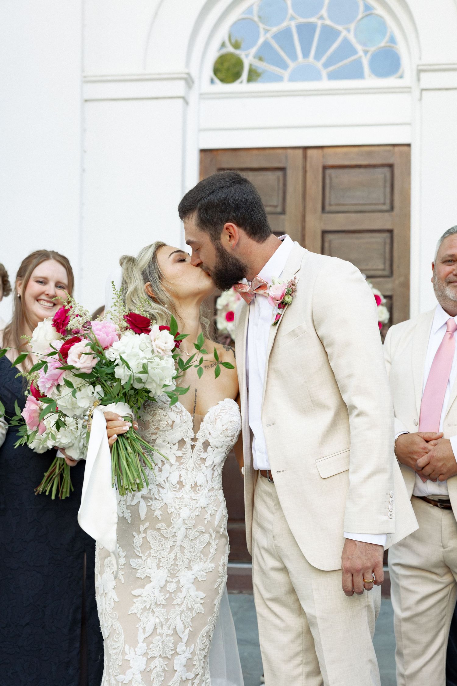 Bride and groom share a first kiss outside white church, bride holding pink and white bouquet in lace wedding gown.