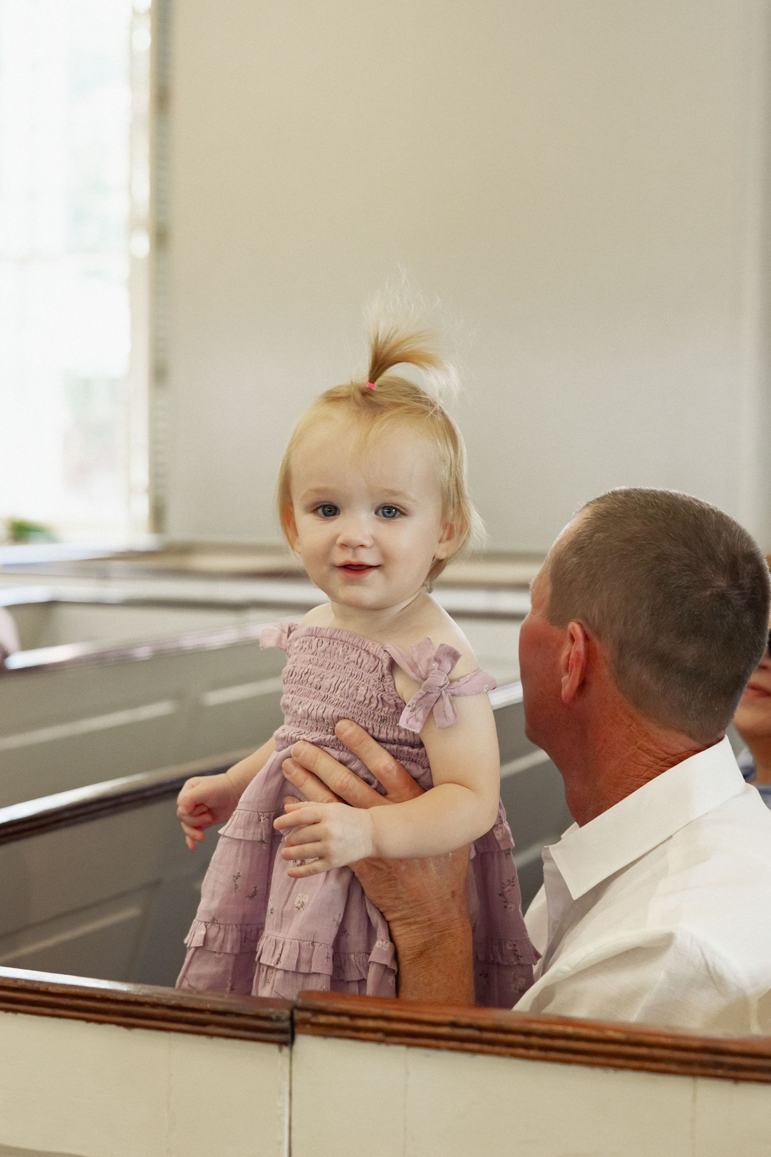 A smiling baby girl in a pink dress being held up by a man in a white shirt inside a church.