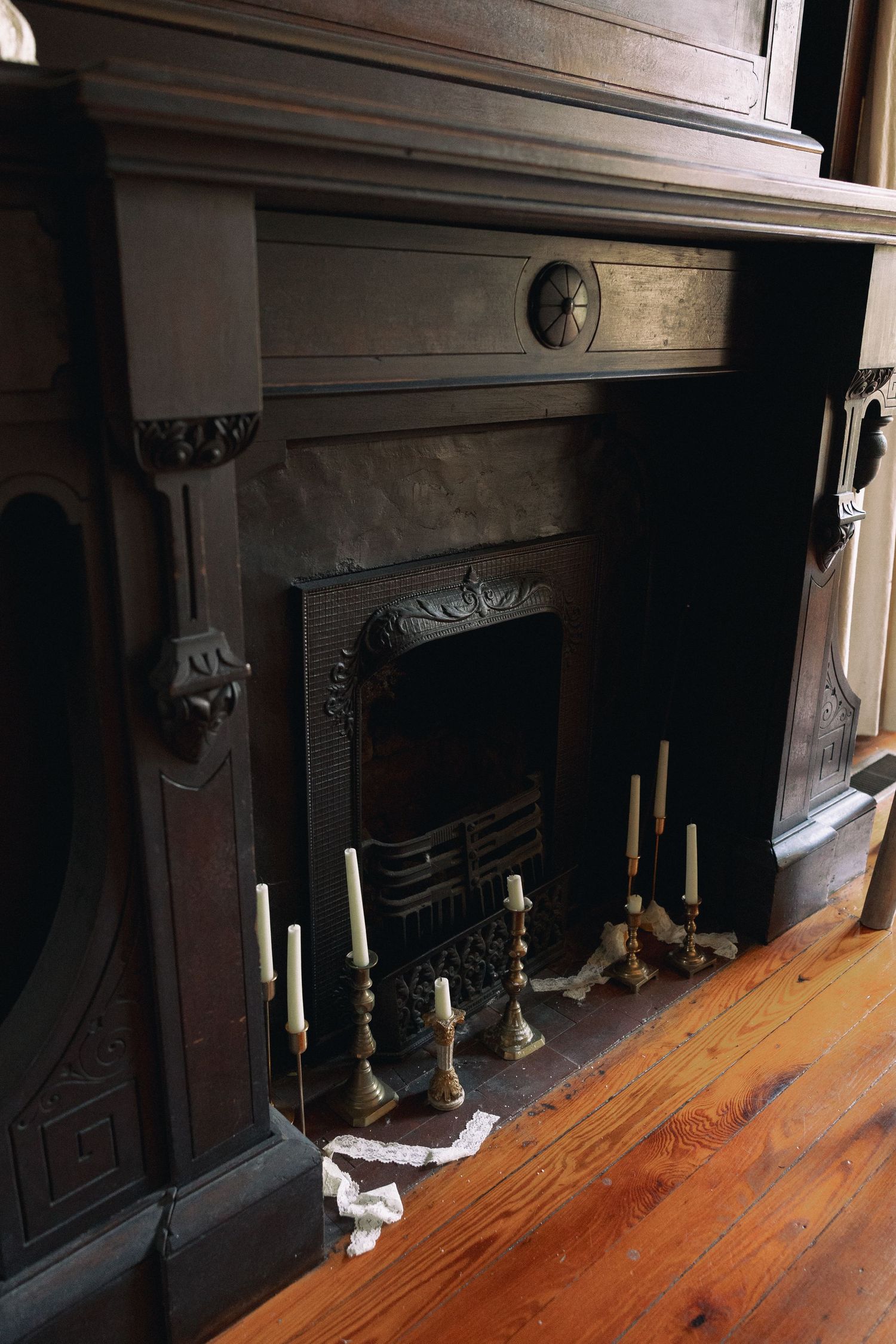Dark ornate Victorian fireplace with decorative carved wood mantel, iron grate, and elegant candlesticks on hardwood floor.