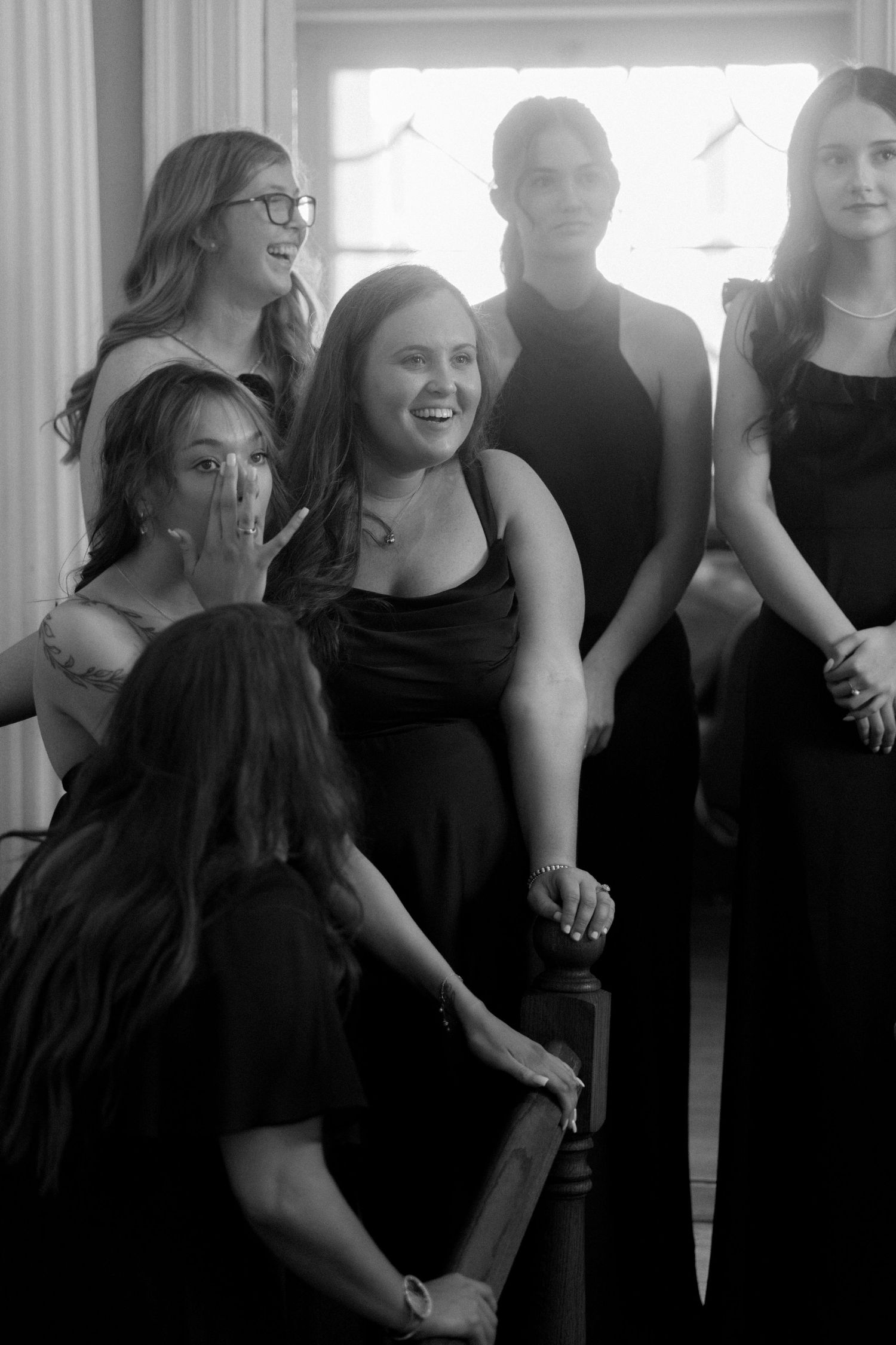 Black and white photo of six young women in elegant black dresses laughing and posing together at a formal event.