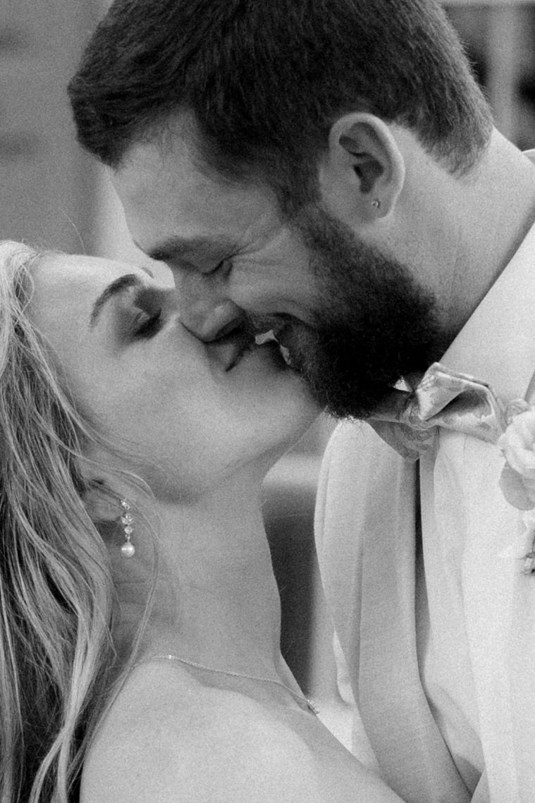 Black and white photo of a couple sharing a romantic kiss, bride in white dress with pearl earrings, groom with beard.
