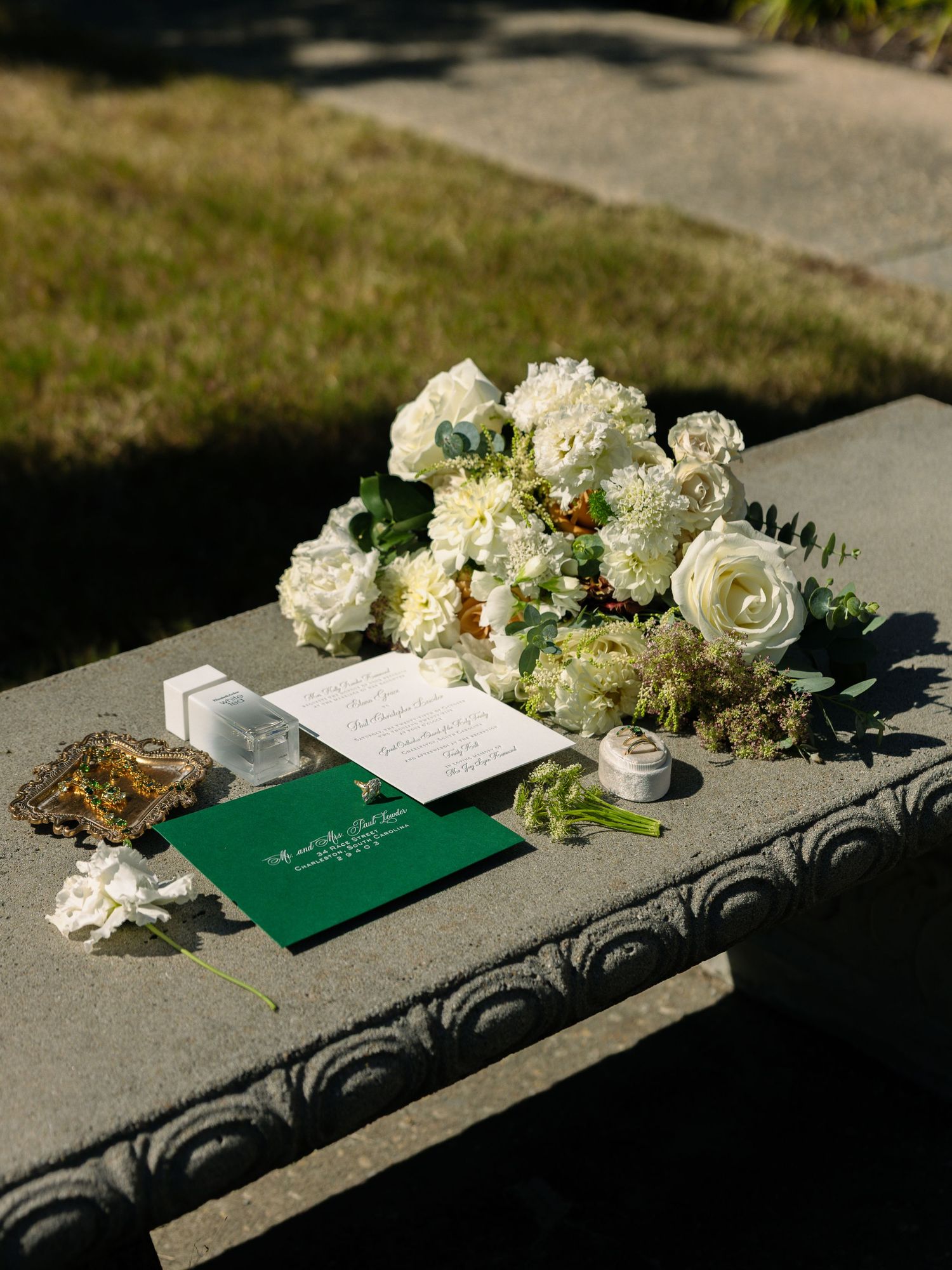 White floral wedding bouquet with roses and hydrangeas beside green invitation on stone bench.
