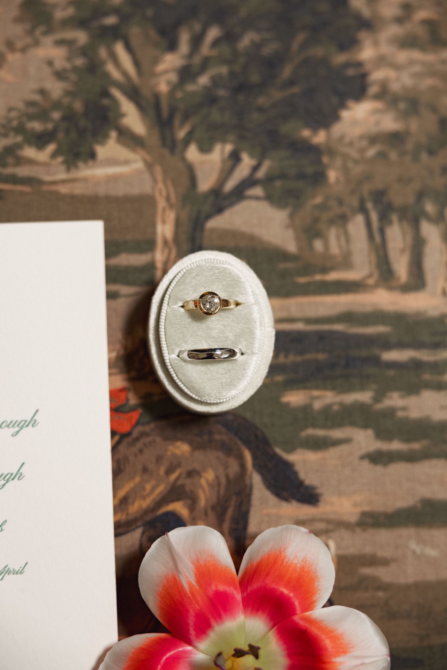 Two wedding rings displayed in oval velvet ring box beside floral invitation and pink tulip.