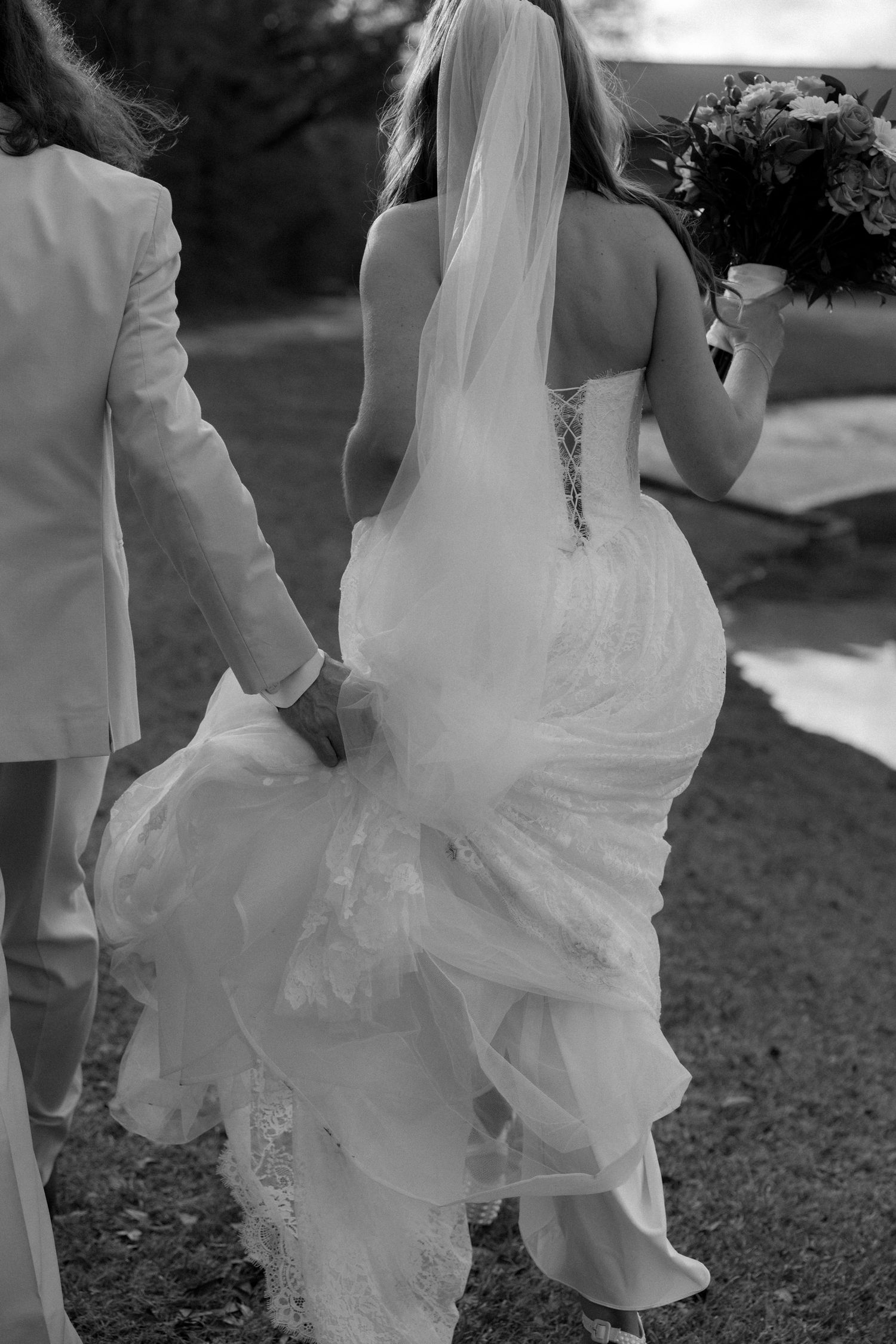 Black and white photo of bride and groom walking hand in hand, bride in lace gown with long veil and bouquet.