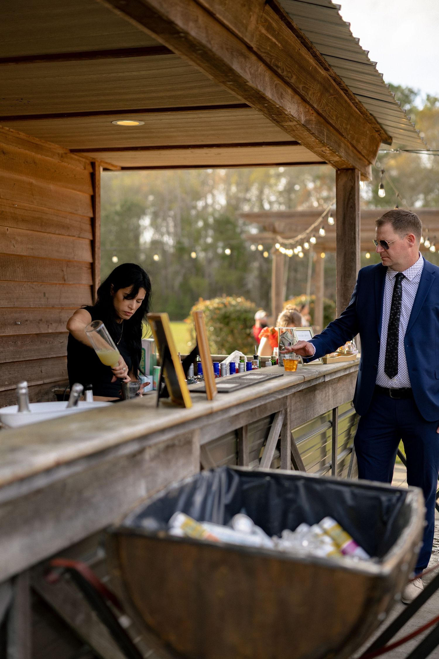 Bartender serving drinks at rustic outdoor wedding bar with string lights and barrel beer cooler.