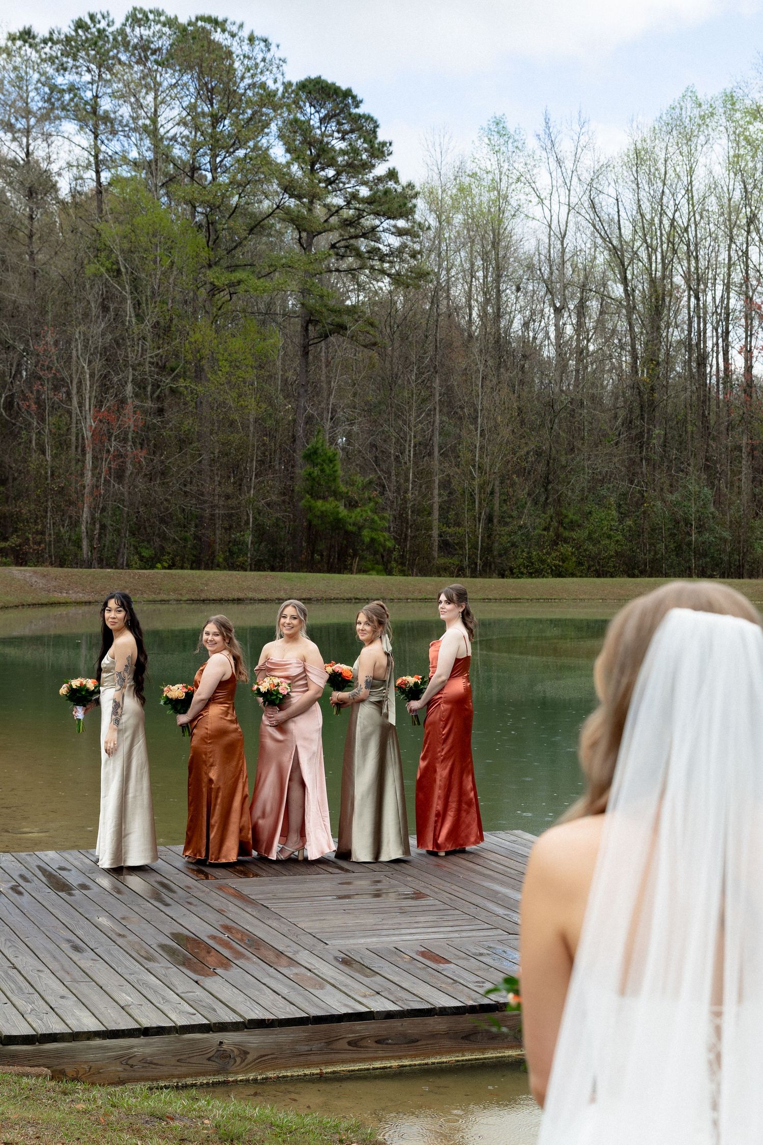 Bridesmaids in rust and champagne satin gowns on dock react to bride's first look reveal.
