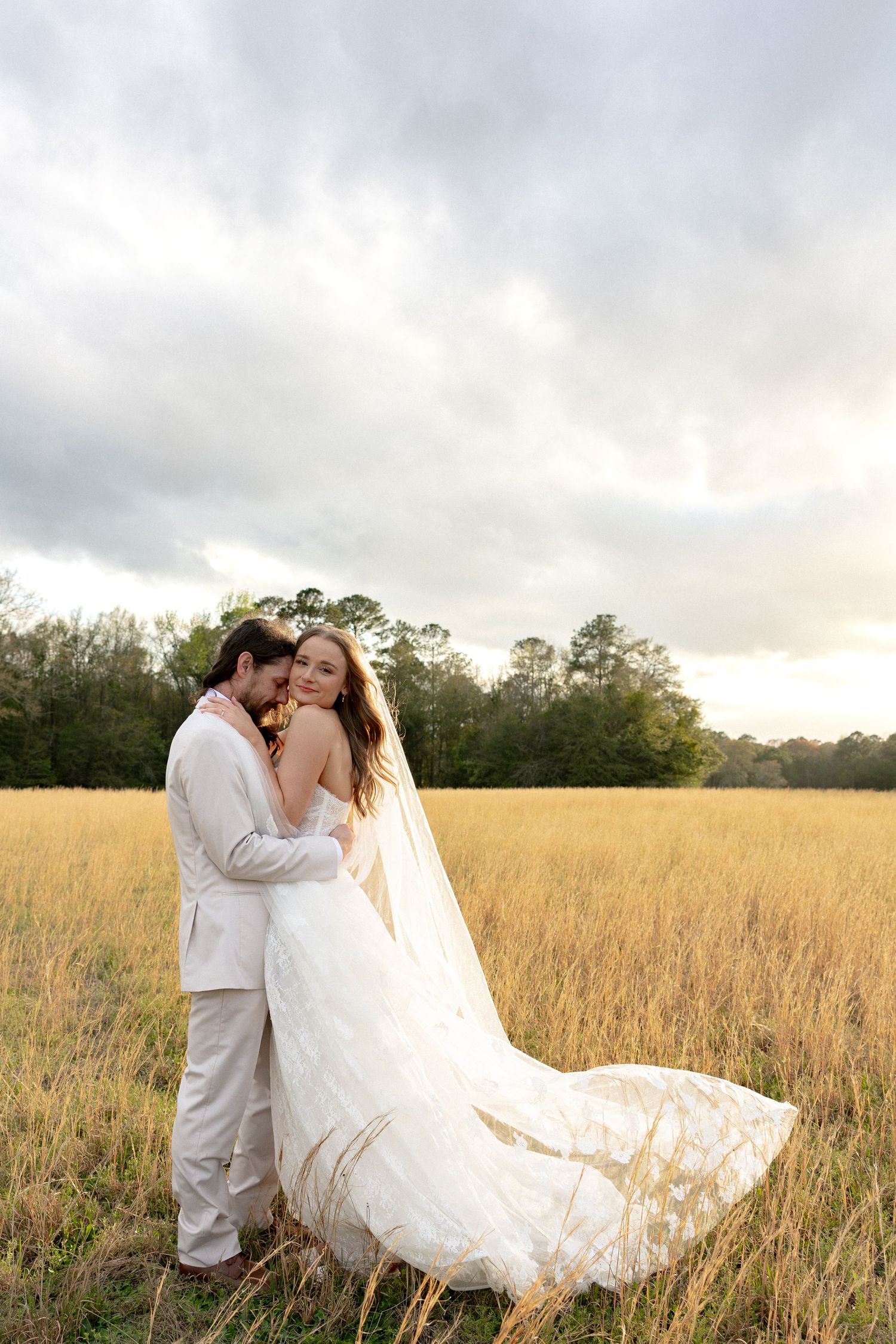 Bride and groom embrace in golden field, her lace gown and veil flowing in the wind.