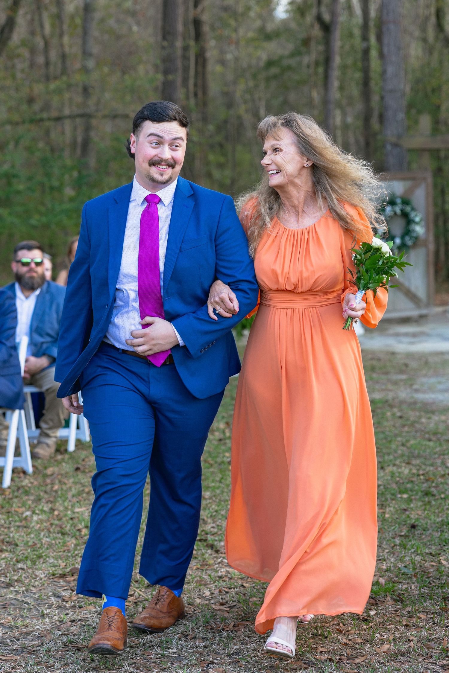 A smiling couple walks down the aisle at an outdoor wedding, man in blue suit with pink tie, woman in orange gown holding bouquet.