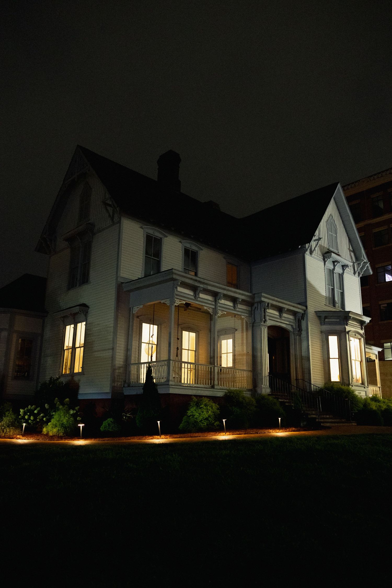 Historic white Victorian house illuminated at night with glowing windows and landscape lighting on dark lawn.
