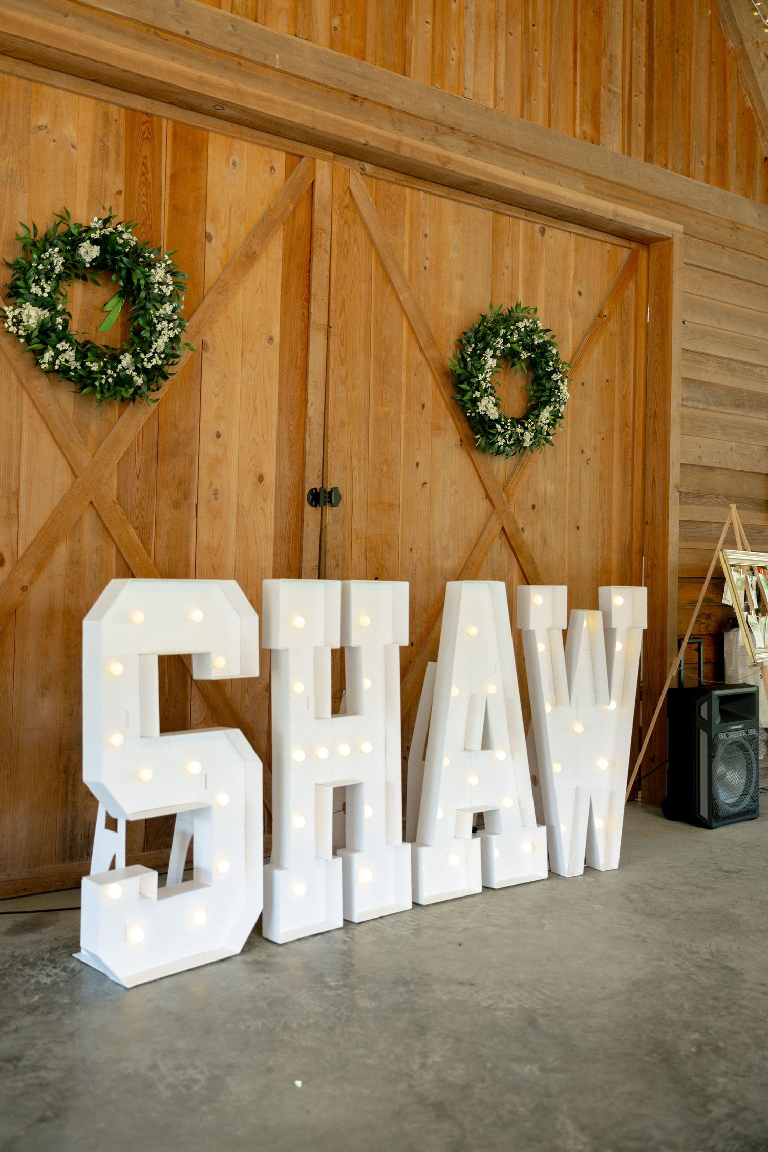 Large illuminated marquee letters spelling SHAW displayed in a rustic wooden barn wedding venue.