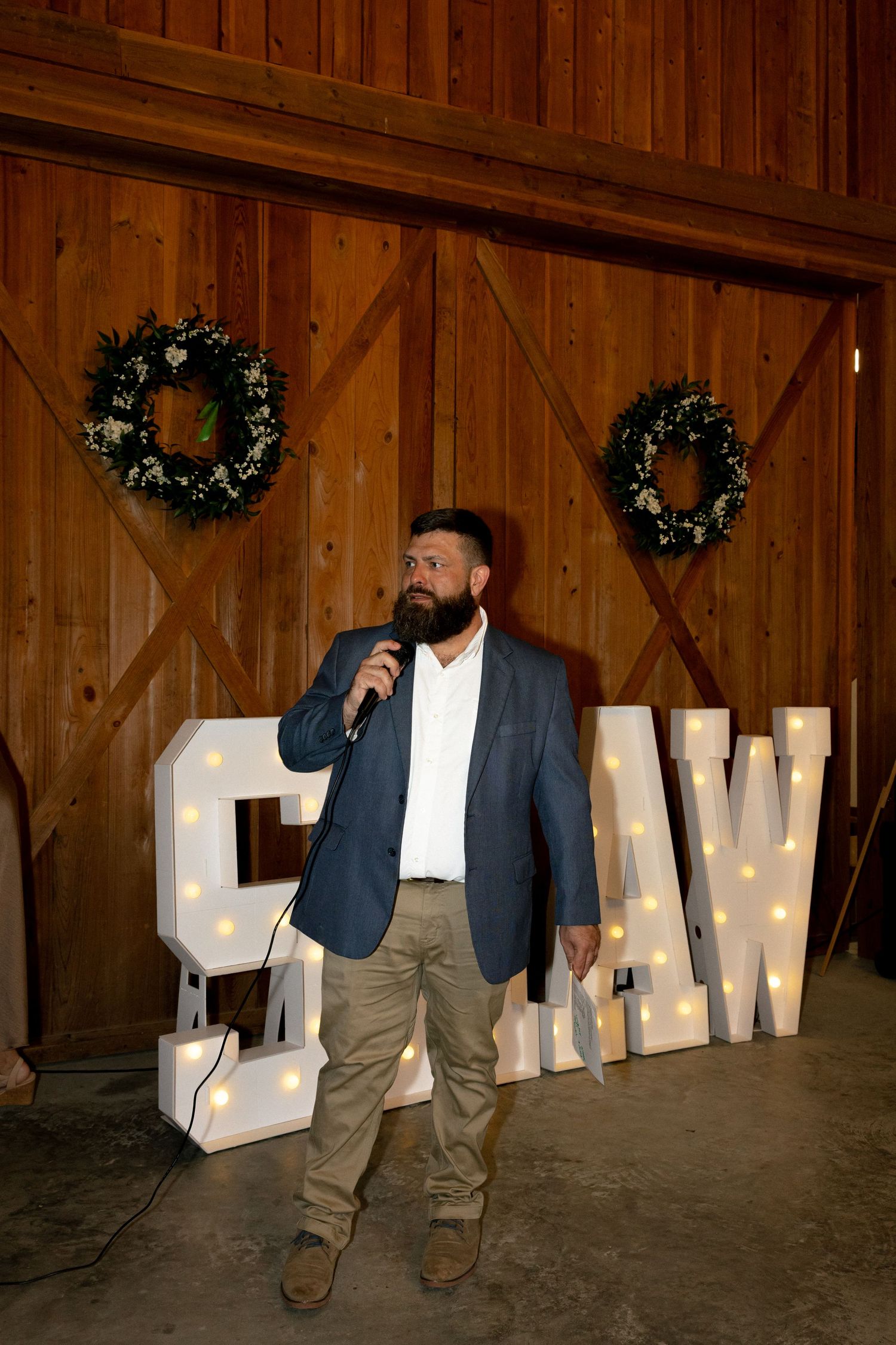 Bearded man in navy blazer giving speech at rustic barn wedding reception with illuminated marquee letters.
