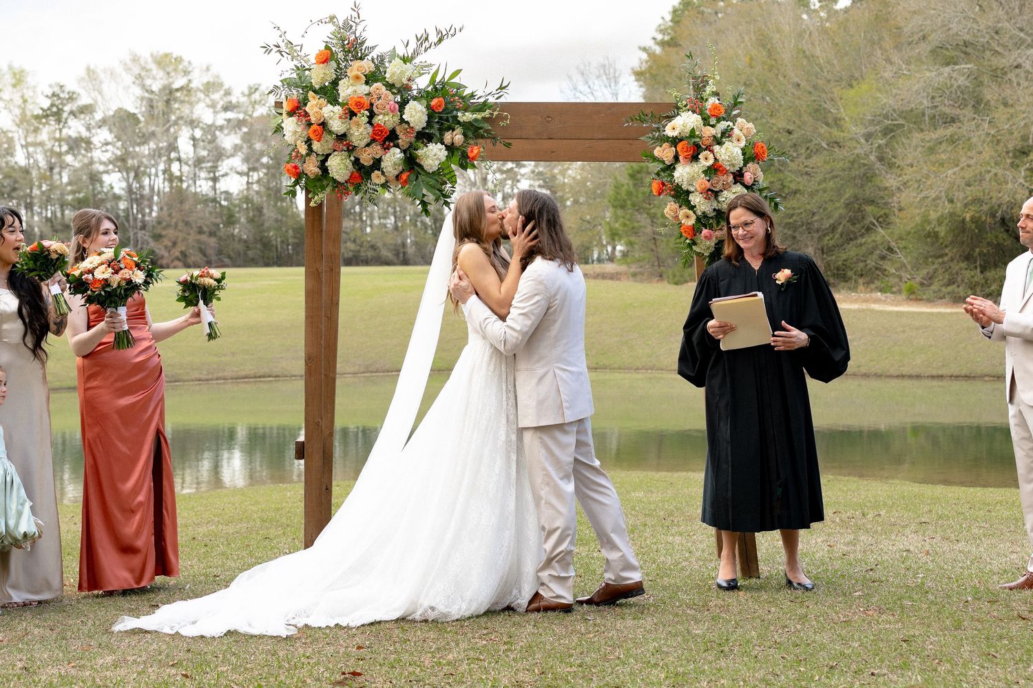 Bride and groom sharing first kiss at outdoor wedding ceremony beneath wooden arch with floral arrangements.