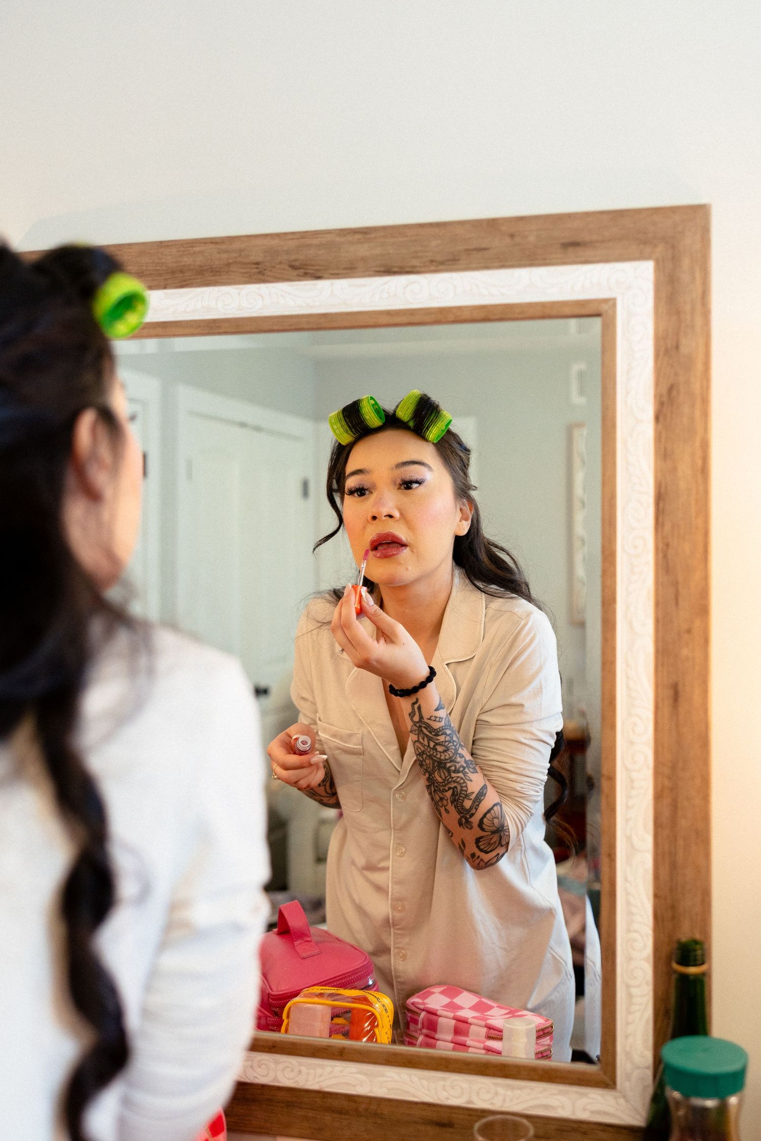 Bride with green hair rollers applying lipstick at a wooden vanity mirror.