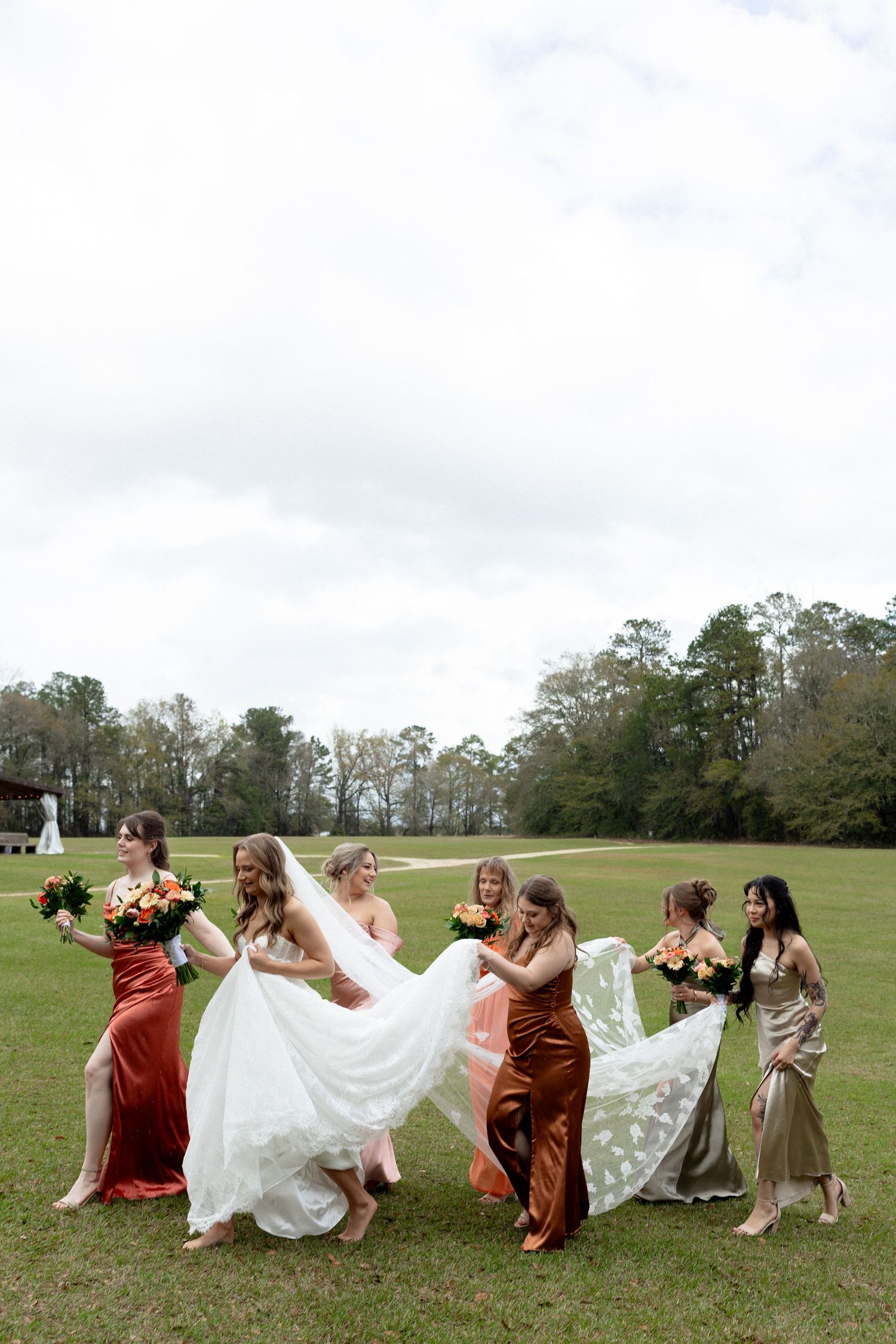Six bridesmaids in rust and gold velvet gowns carry bride's white wedding dress train across green lawn.