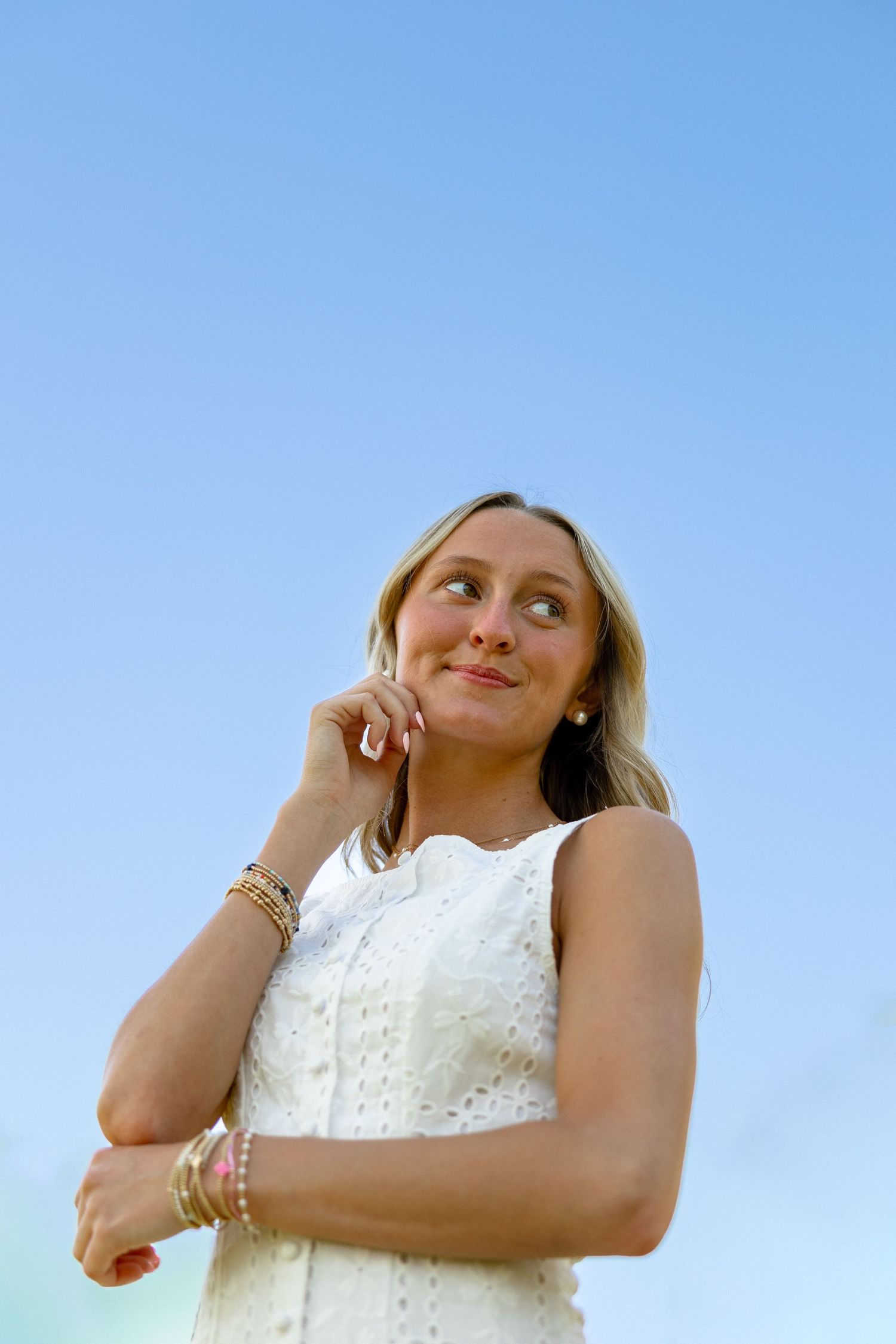 Thoughtful young blonde woman in a white dress posing against a clear blue sky background.
