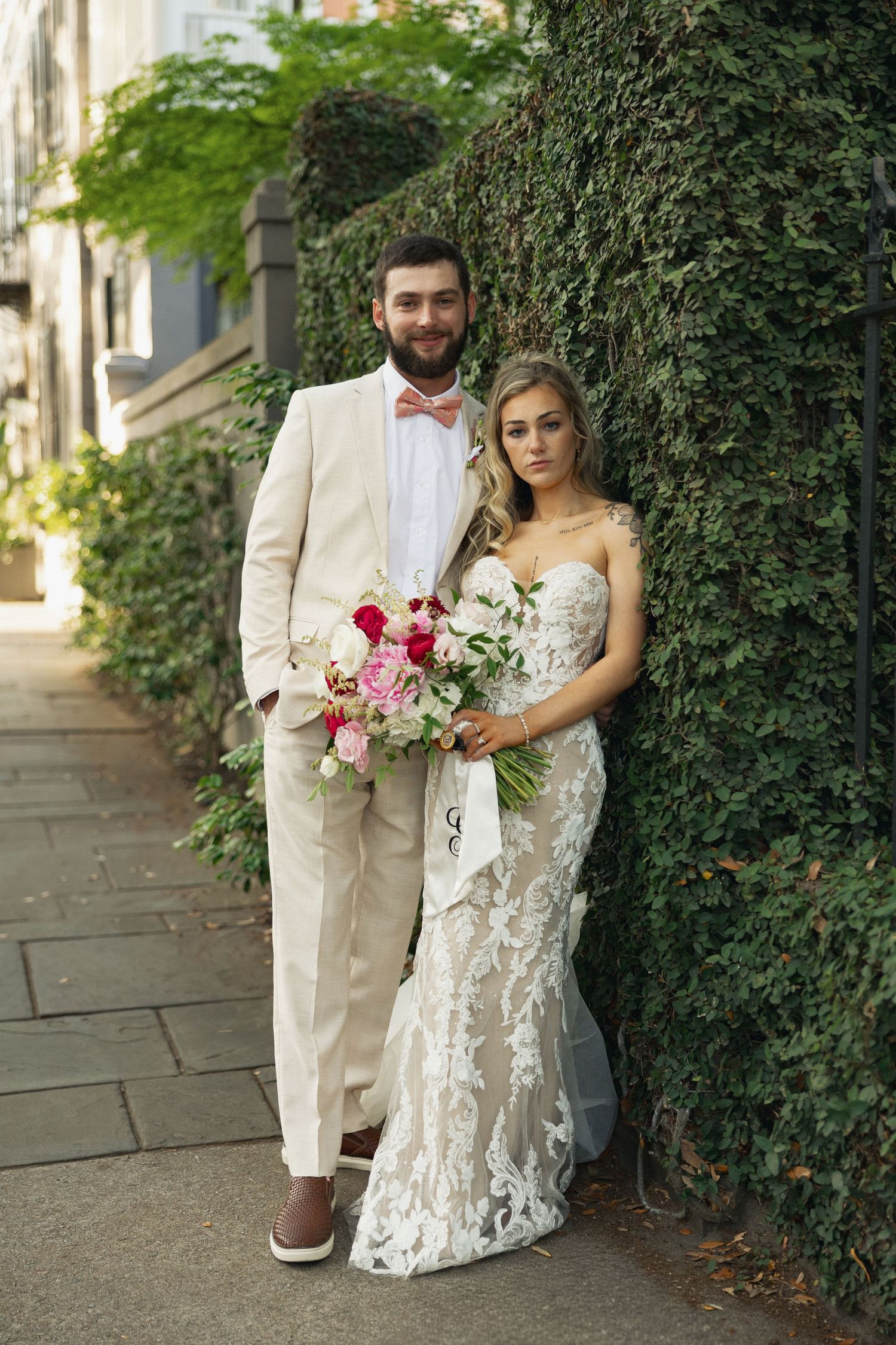 Bride in lace mermaid gown holds pink bouquet beside groom in cream suit with pink bow tie on ivy-covered wall.