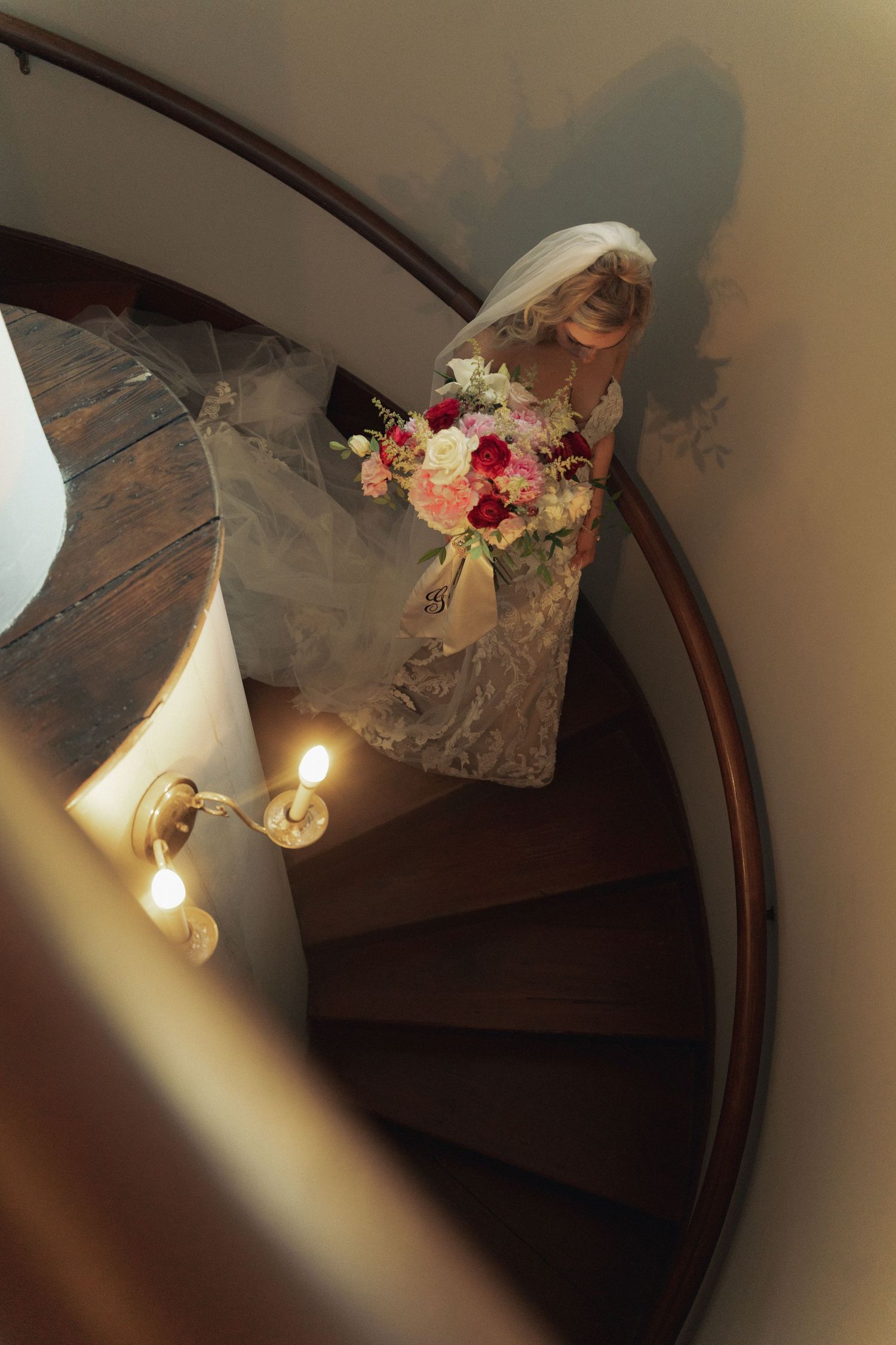 Bride in lace gown holding floral bouquet descends elegant spiral staircase with warm brass lighting.