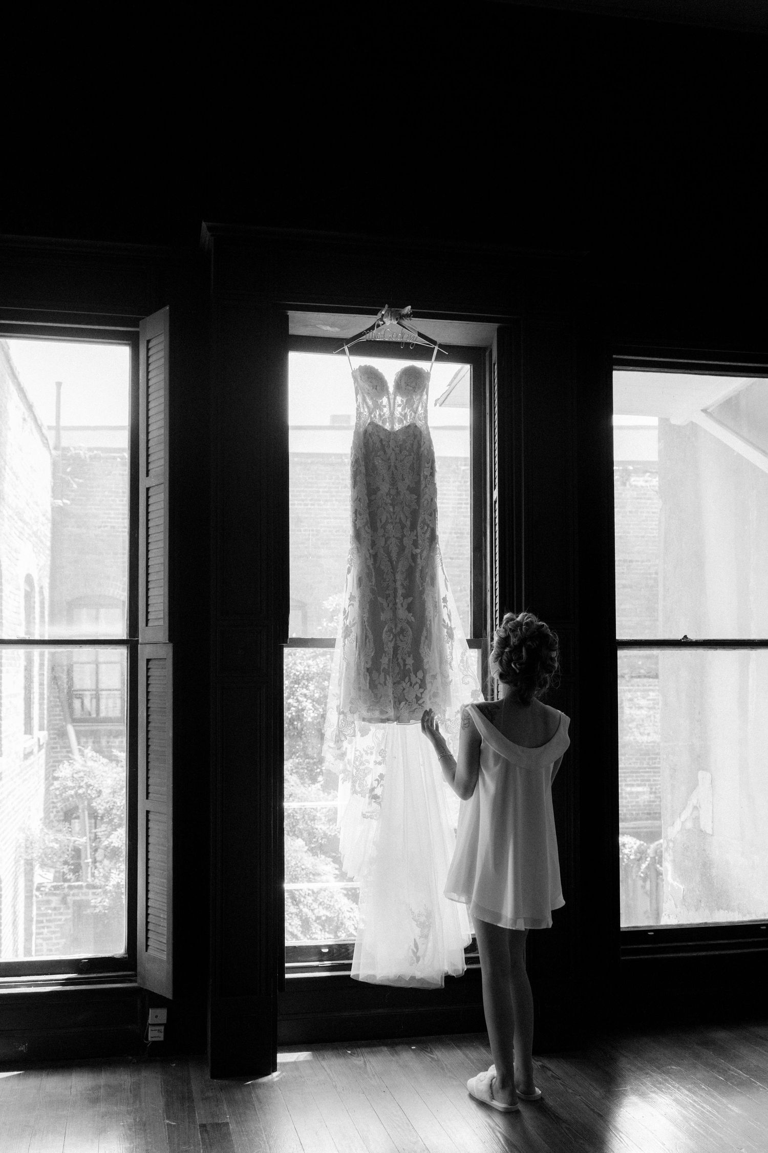 Black and white photo of a young girl in white dress admiring a wedding gown hanging in a sunlit window.