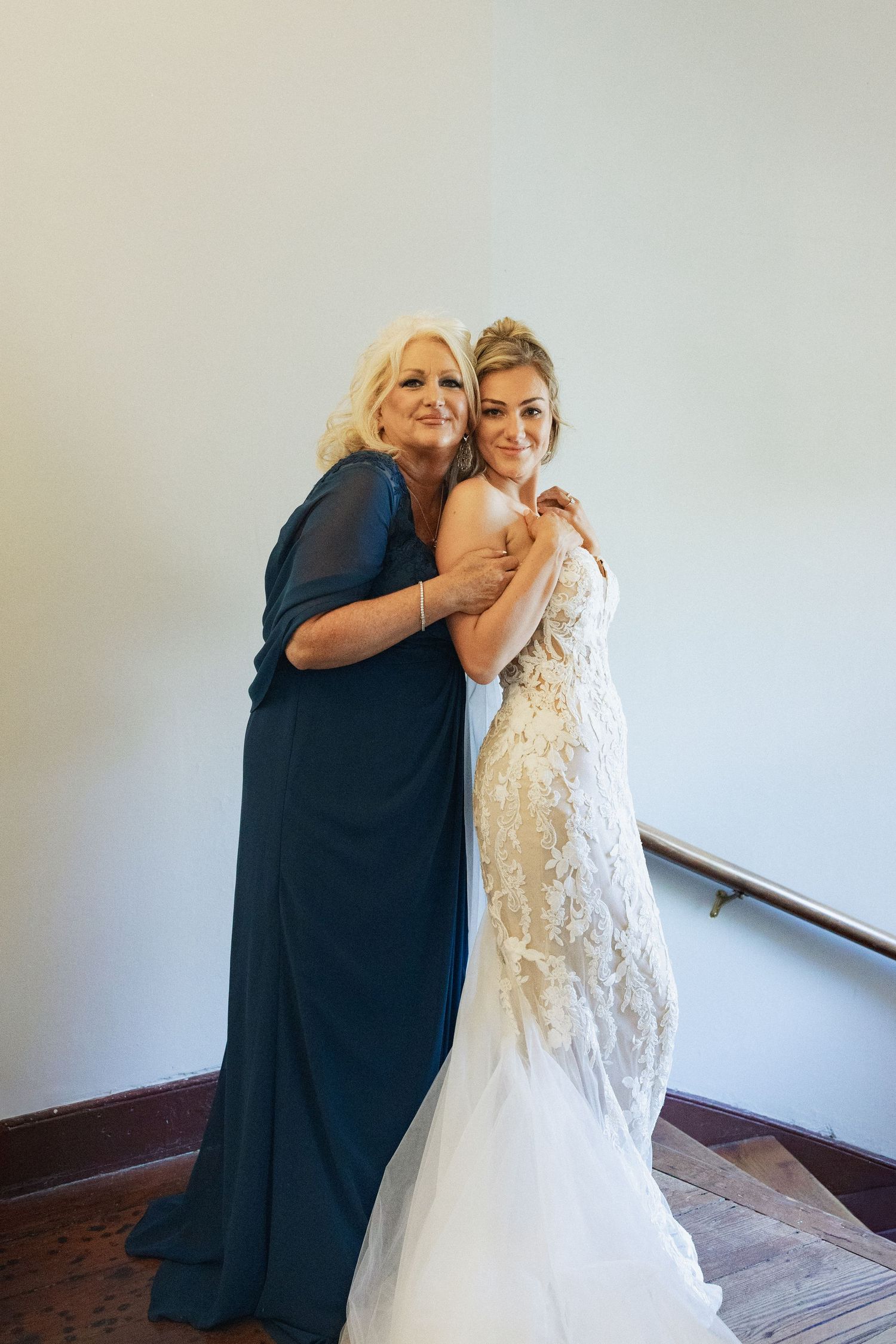 A bride in a lace wedding gown shares a warm embrace with an older woman in a teal formal dress.