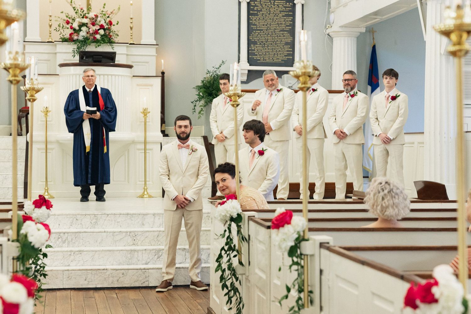 Groom and groomsmen in cream suits stand at church altar during wedding ceremony with officiant in blue robe.