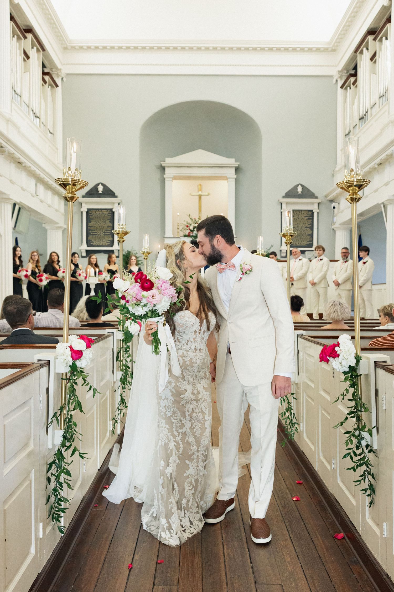 Newlyweds share first kiss in church aisle surrounded by red and white floral decorations.