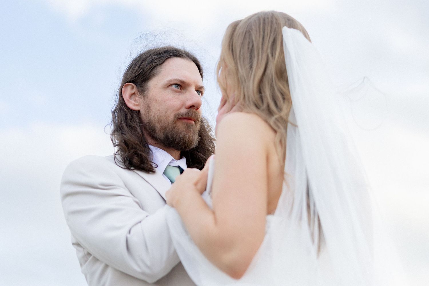 Bearded groom in tan suit gazes lovingly at bride in white veil outdoors.