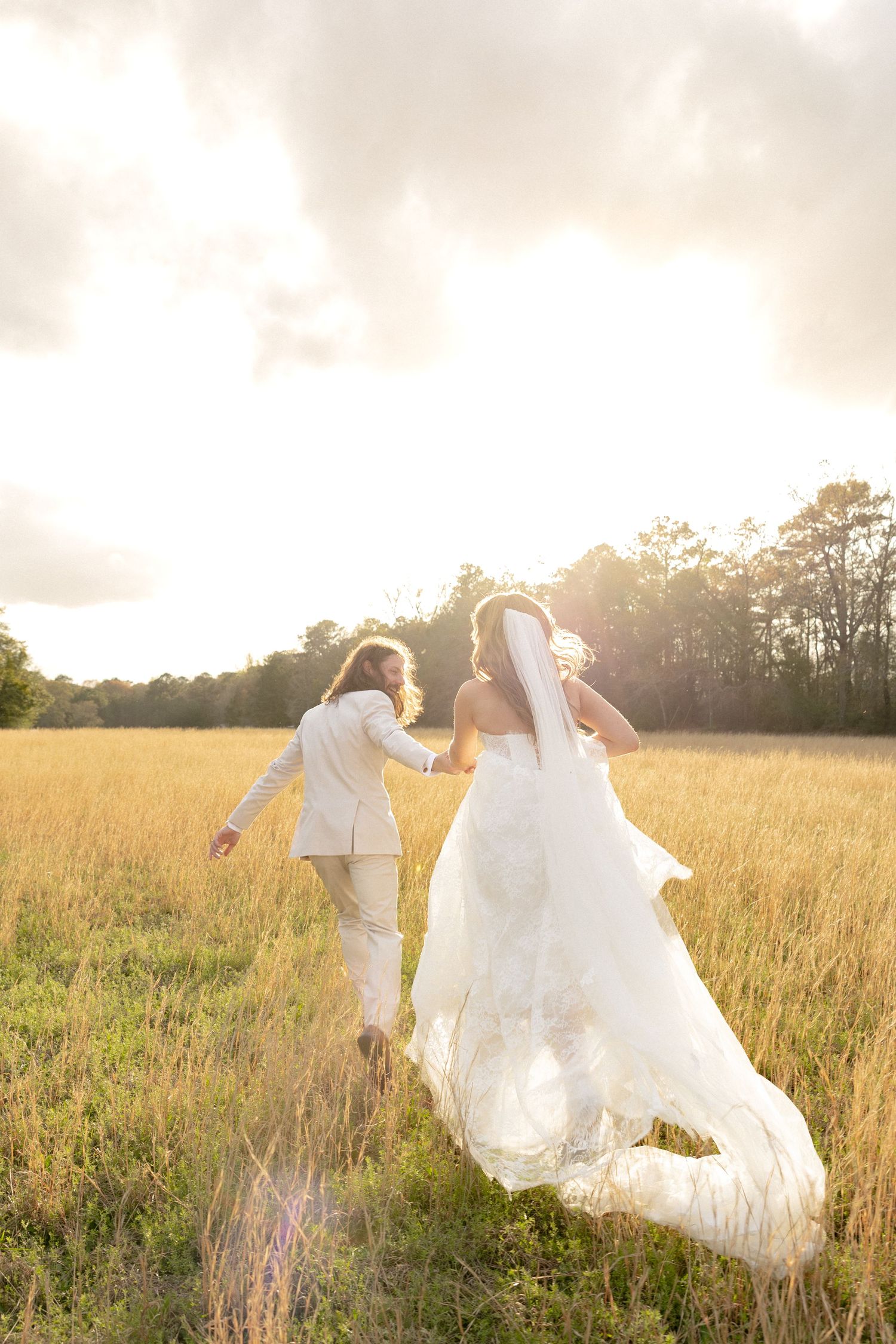 Bride and groom running hand-in-hand through a golden field at sunset on their wedding day.