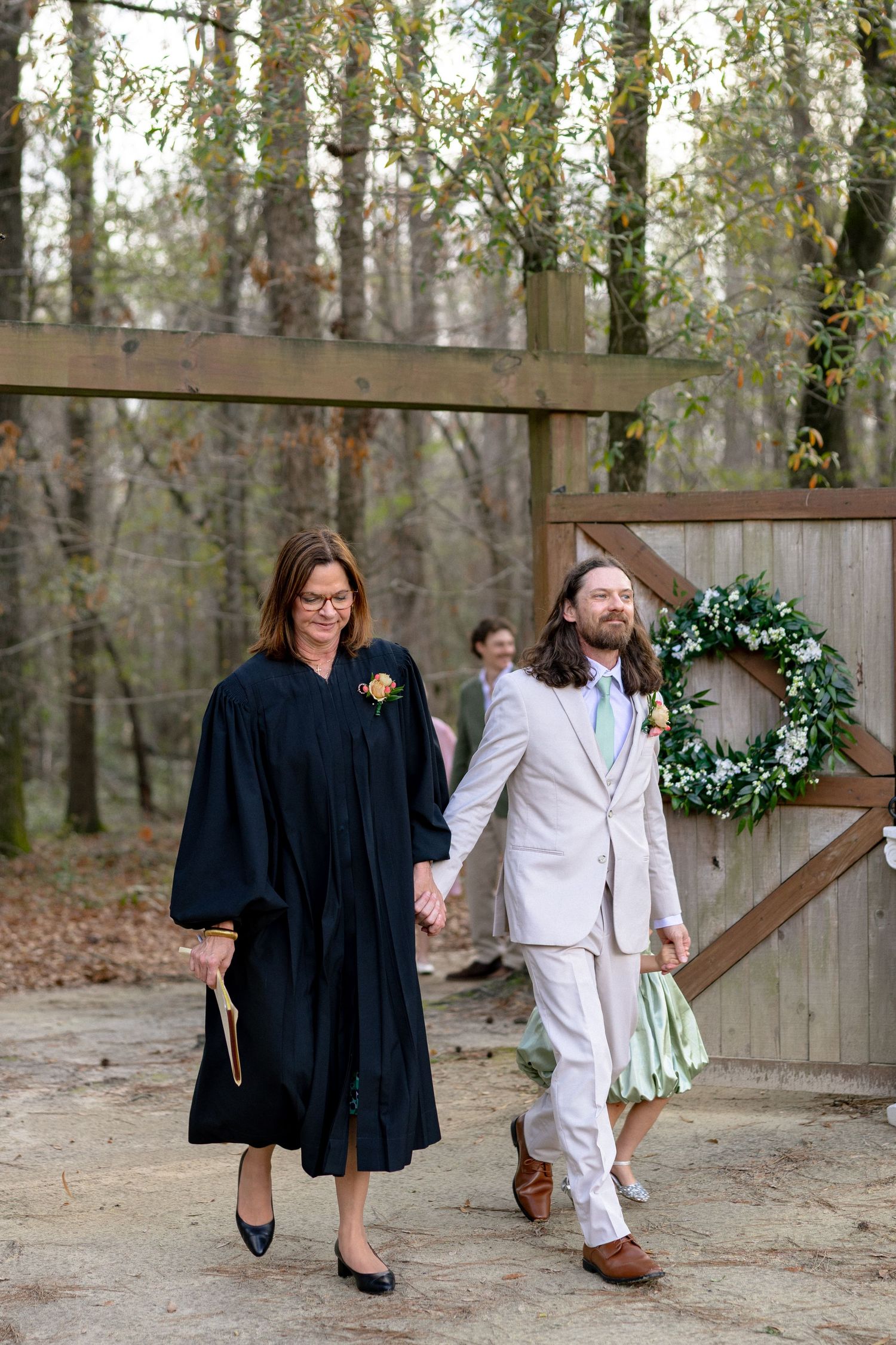 Officiant in black robes walking with the groom in a tan suit through a wooden gate adorned with a wreath.
