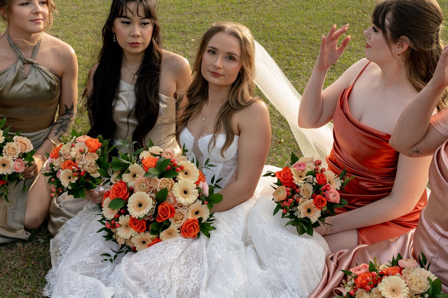 Bride in white gown sits with bridesmaids in coral and gold dresses holding bouquets of orange and white flowers outdoors.