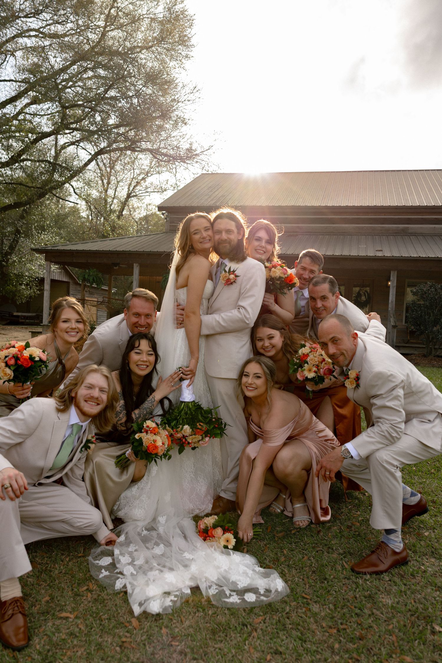 Wedding party surrounds embracing newlyweds in a close group portrait with golden backlight near a barn.