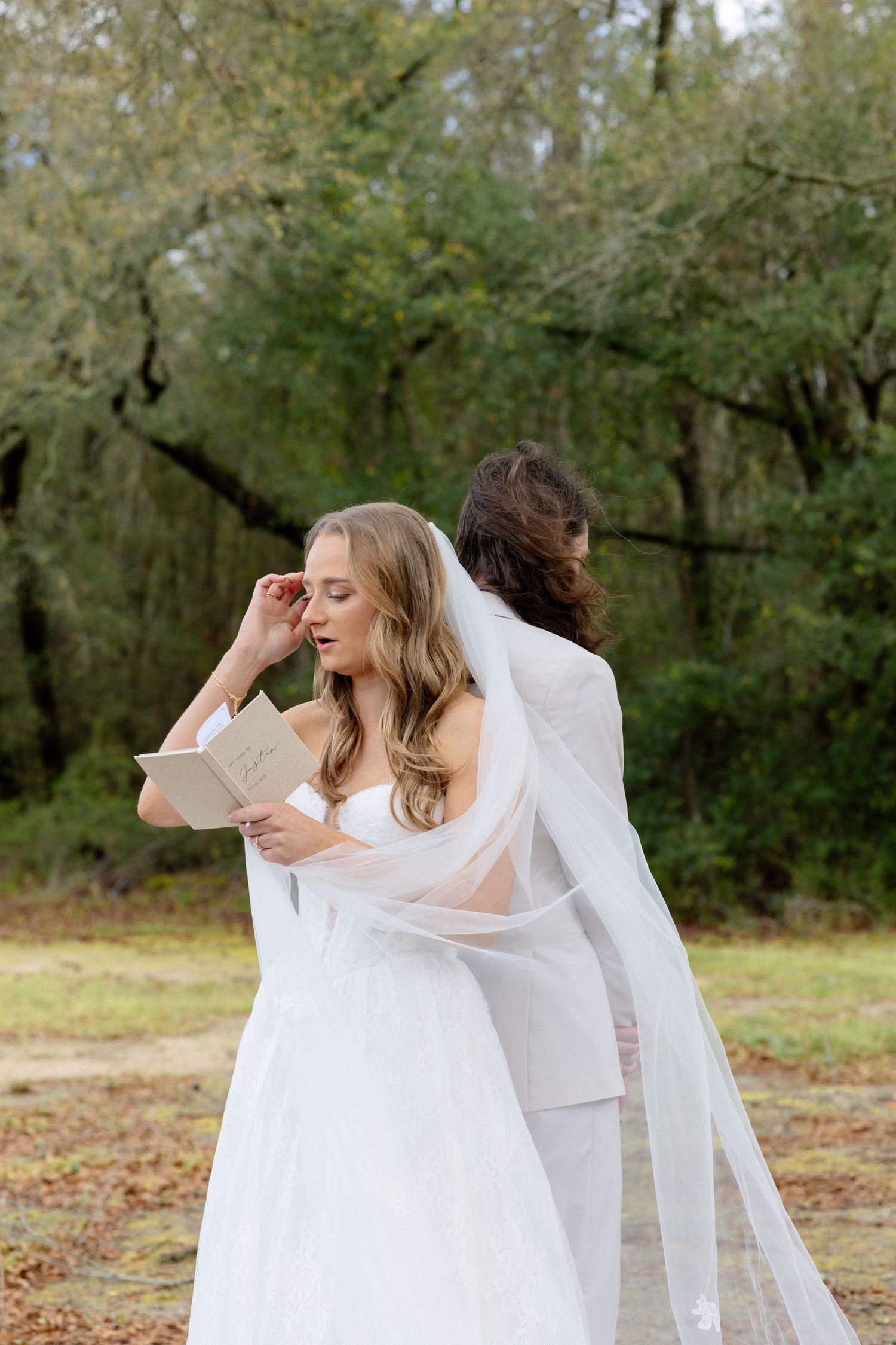 Emotional bride wipes tears reading vow book in white gown and cathedral veil during first look.