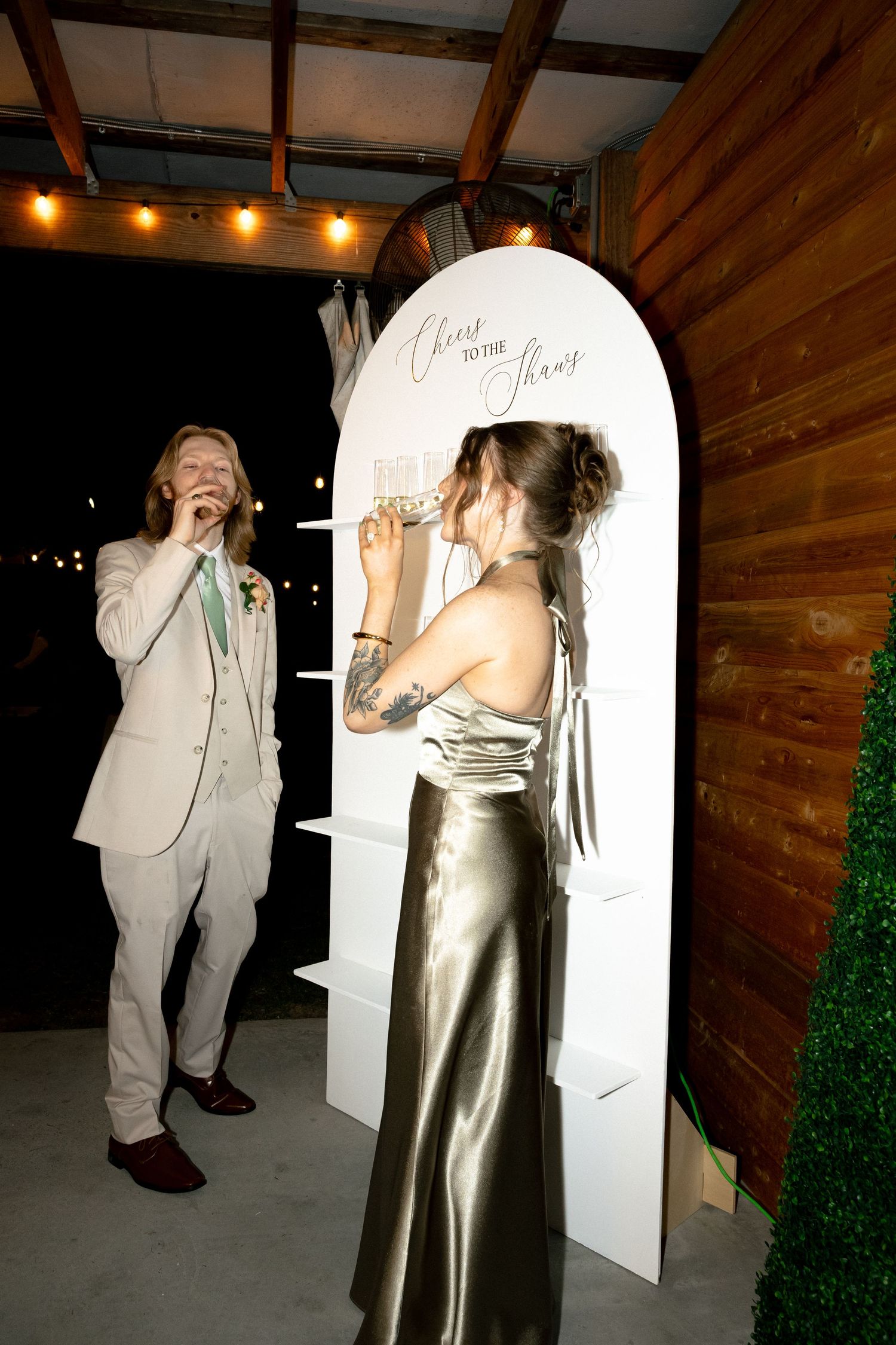 Bride in gold satin gown writes on wedding sign while groom in white suit laughs at rustic barn reception.