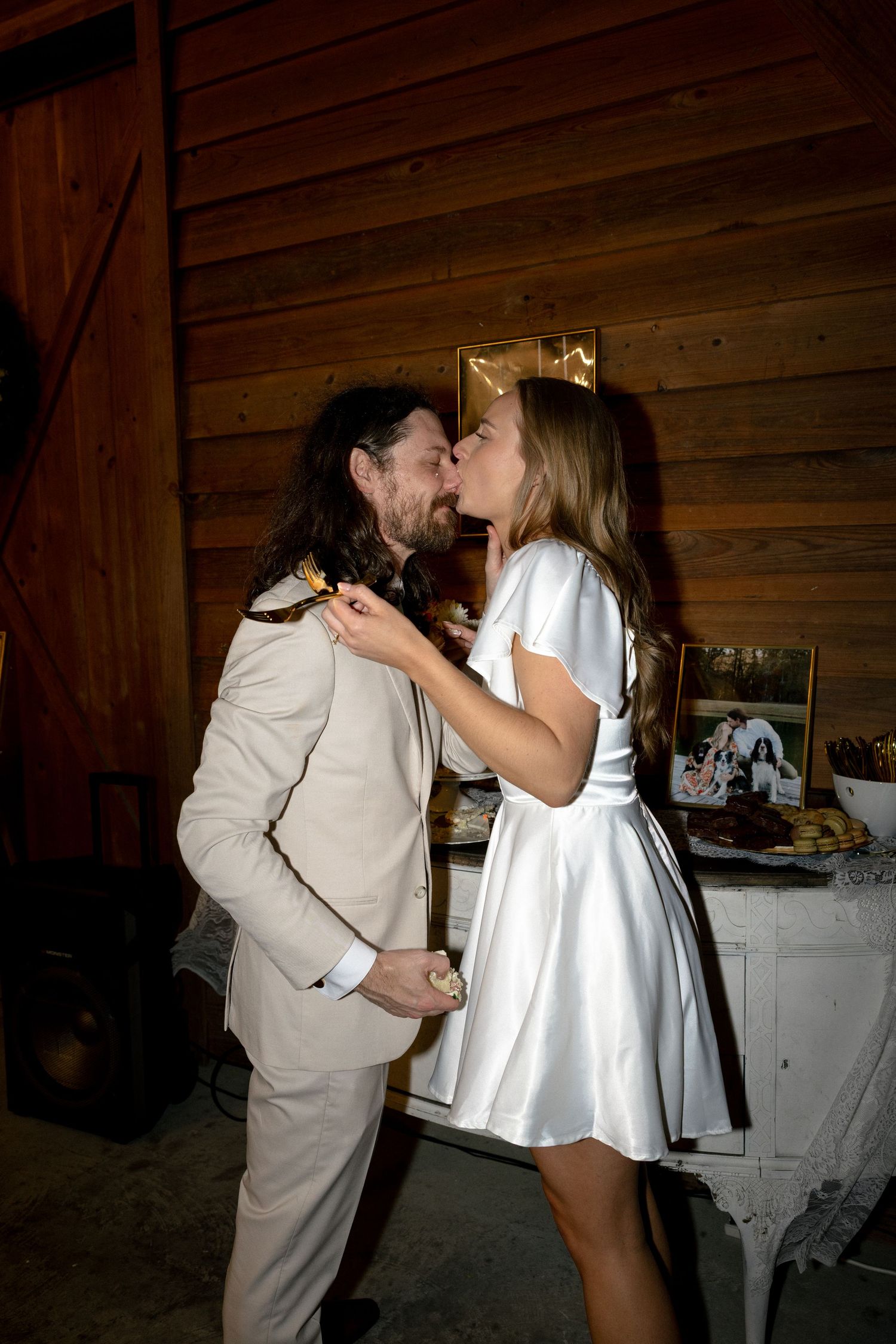 Newlyweds share a romantic kiss at their rustic barn wedding reception near the cake table.