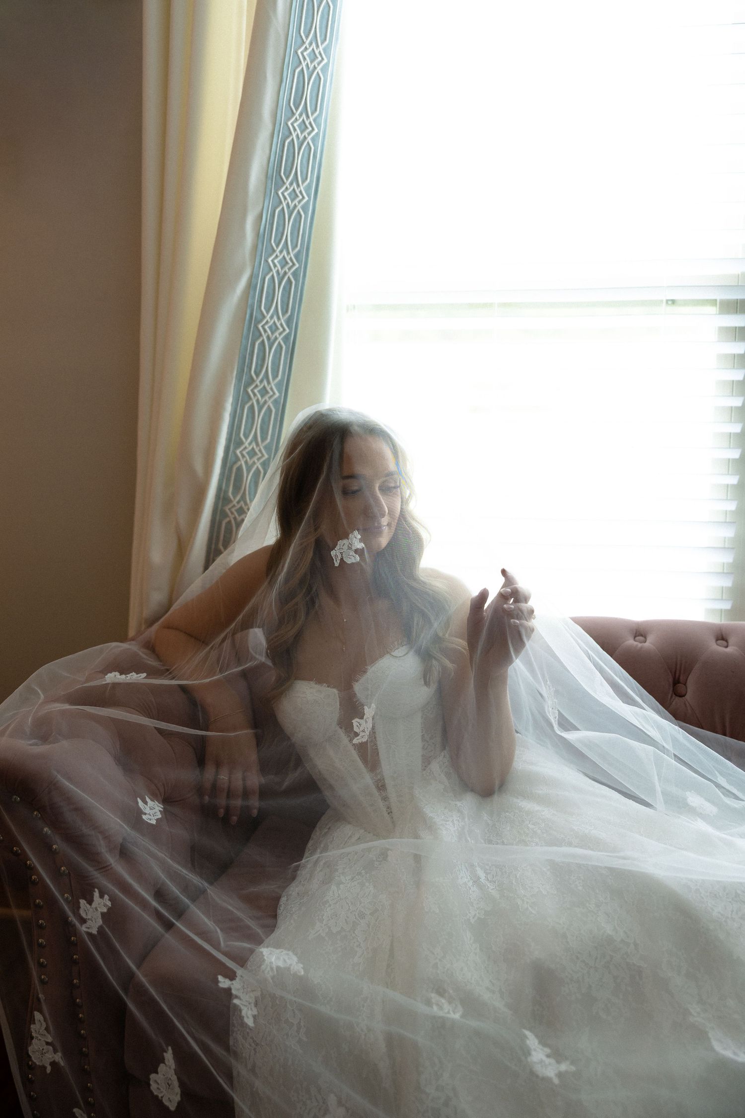 Bride in lace wedding gown covered by floral veil, seated by bright window holding a flower.