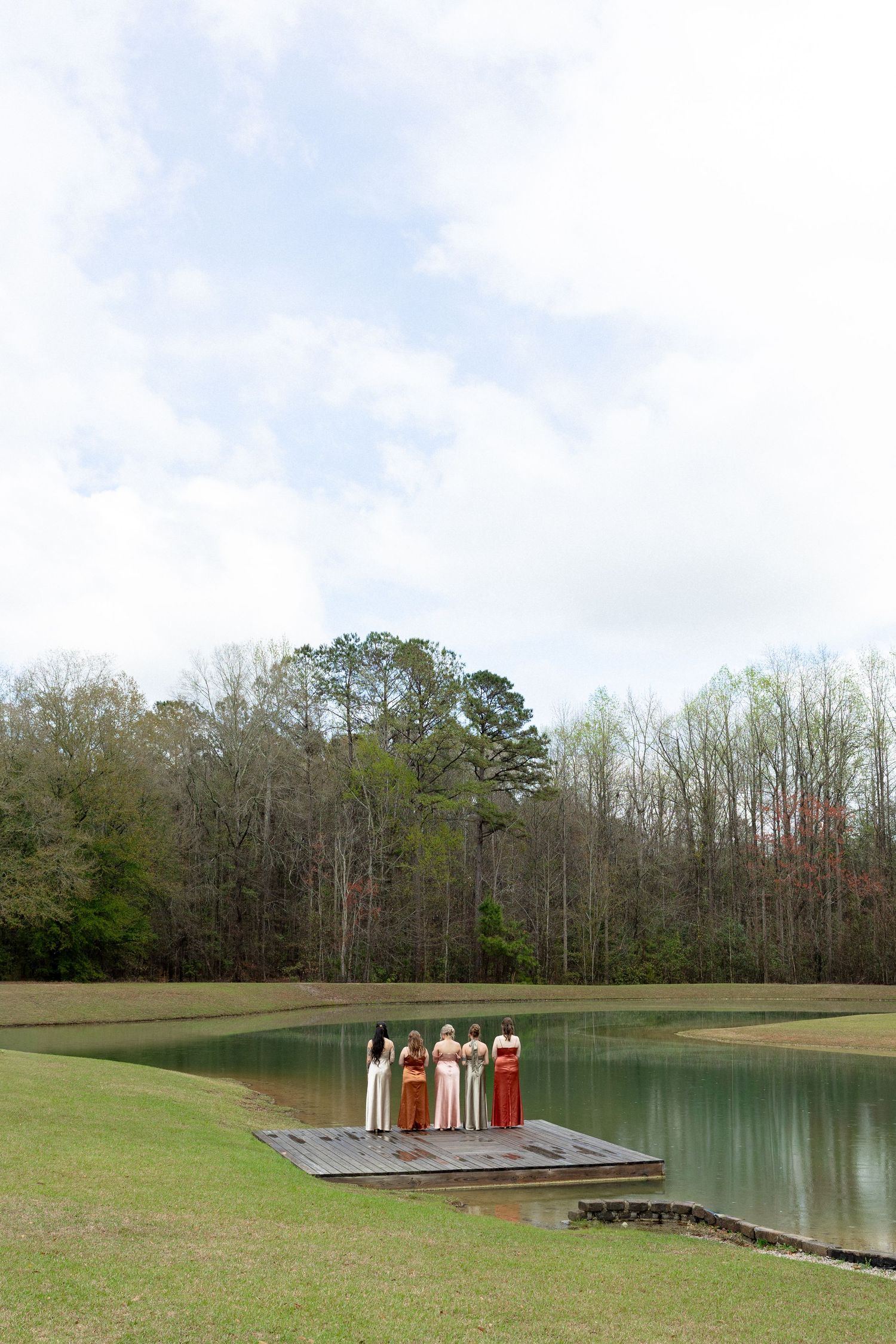 Bridesmaids in mismatched earth-tone gowns standing on a wooden dock beside a serene pond.