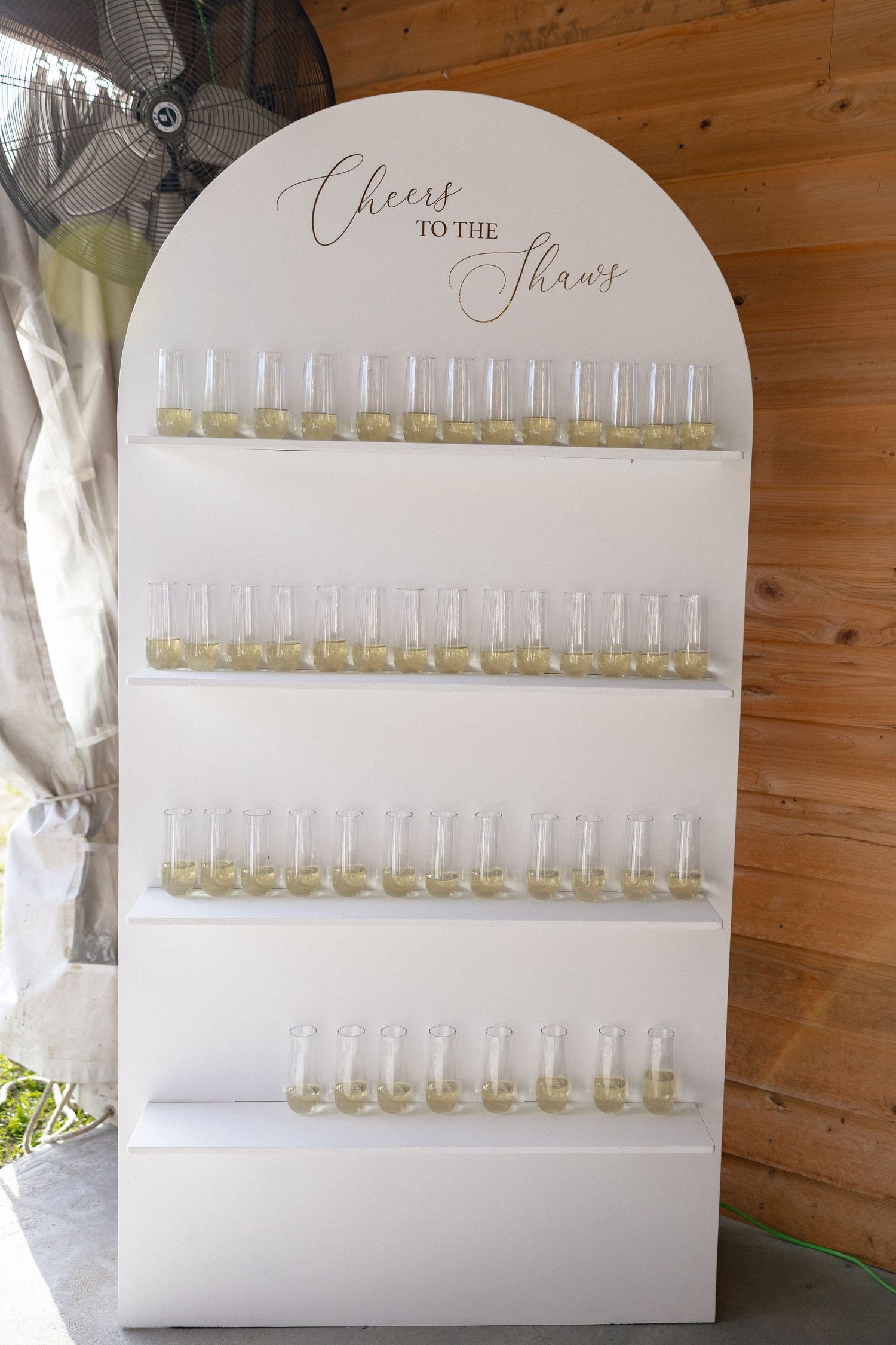 White arched champagne wall display reading Cheers to the Shaws with rows of filled flute glasses.