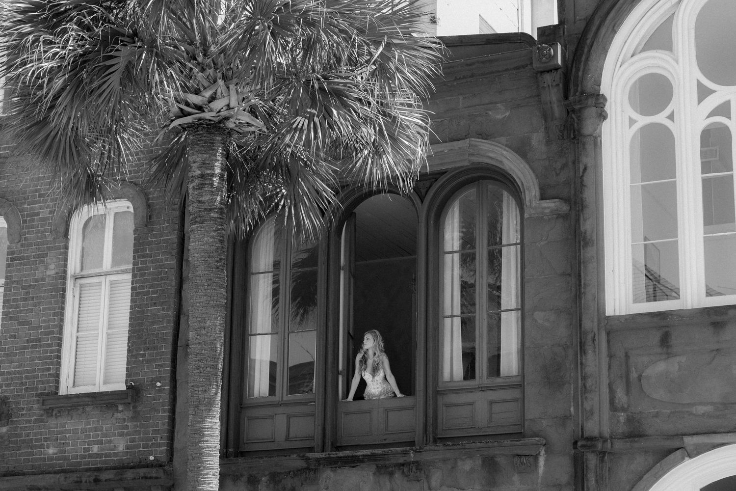 Black and white photo of a woman sitting in an arched window of a historic brick building with a palm tree.