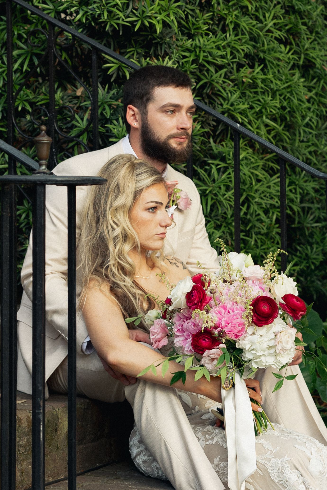 Bride and groom sitting on stone steps surrounded by greenery, bride holding a lush pink and red floral bouquet.