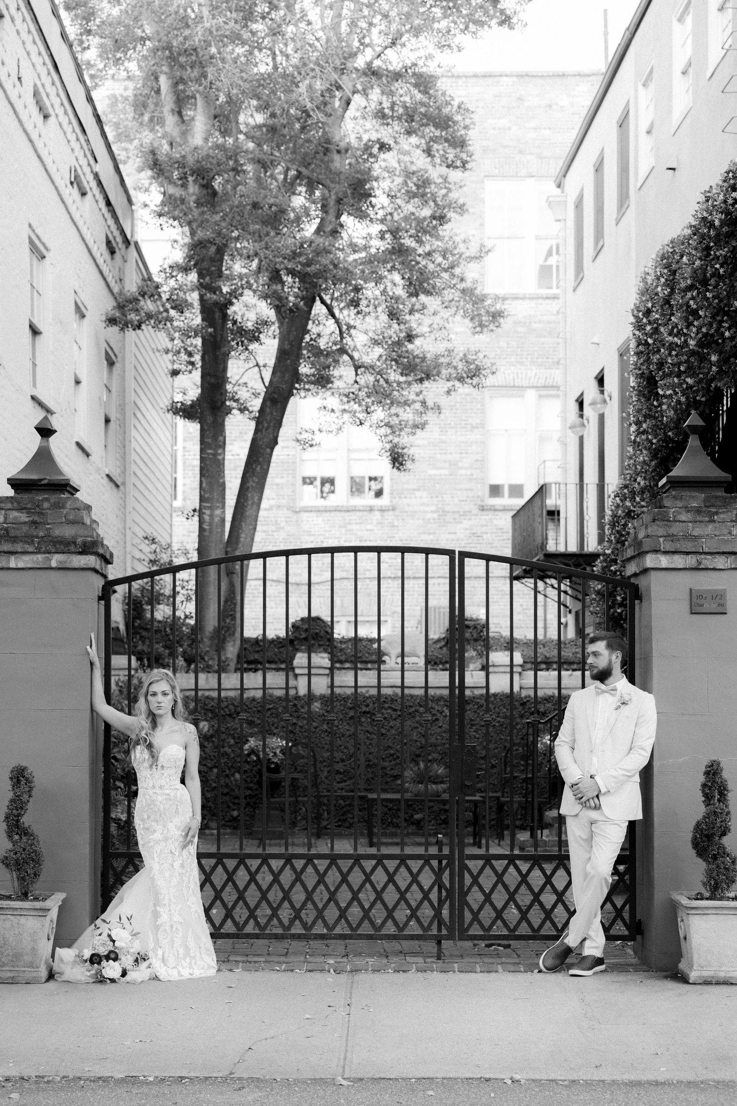 Black and white wedding portrait of bride and groom posing at ornate iron gate on city street.